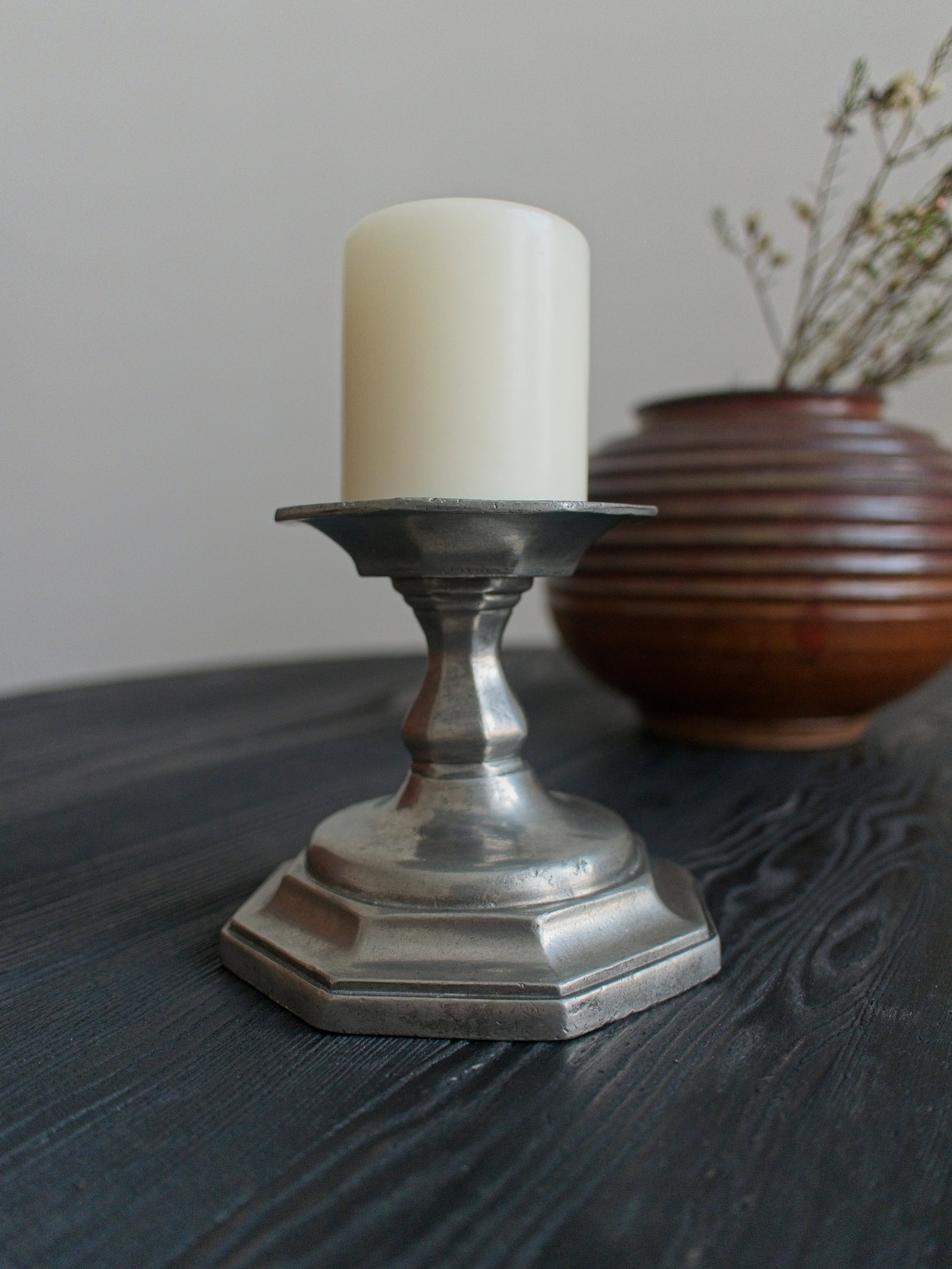 A vintage silver candle holder with a cream candle on a dark surface, with a brown vase and branches in the background.