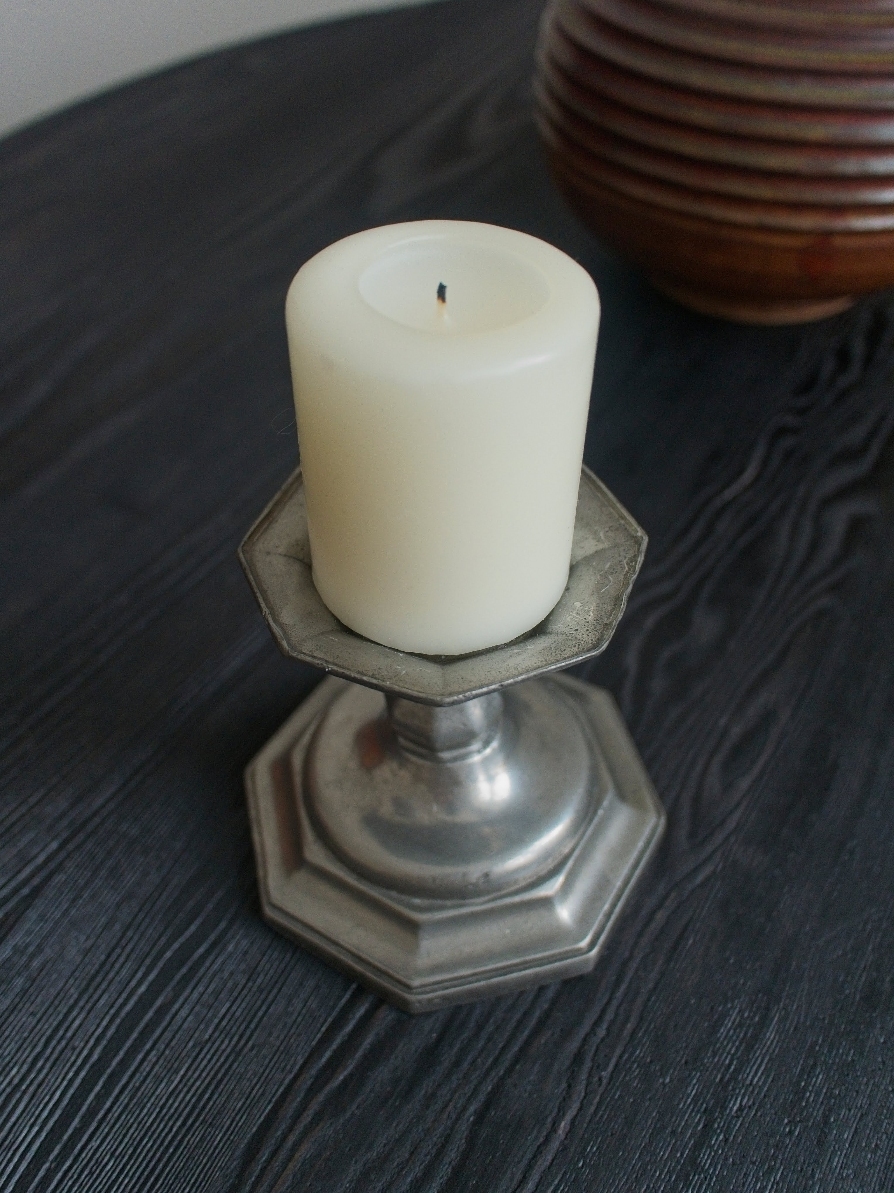 A cream candle in an antique pewter candle holder on a dark surface with a vintage brown textured vase in the background taken from above.