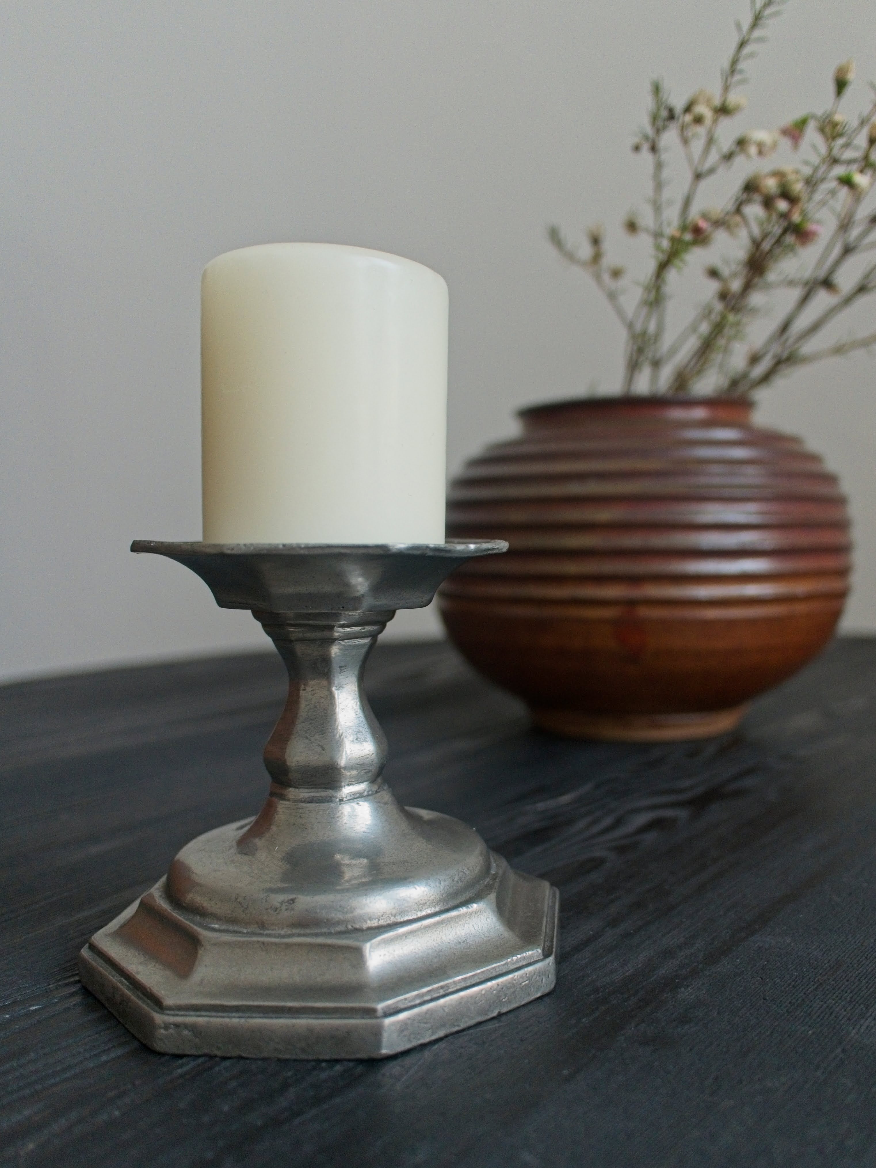 A vintage silver candle holder with a cream candle on a black wood table, next to an art deco ridged vase with dried flowers.