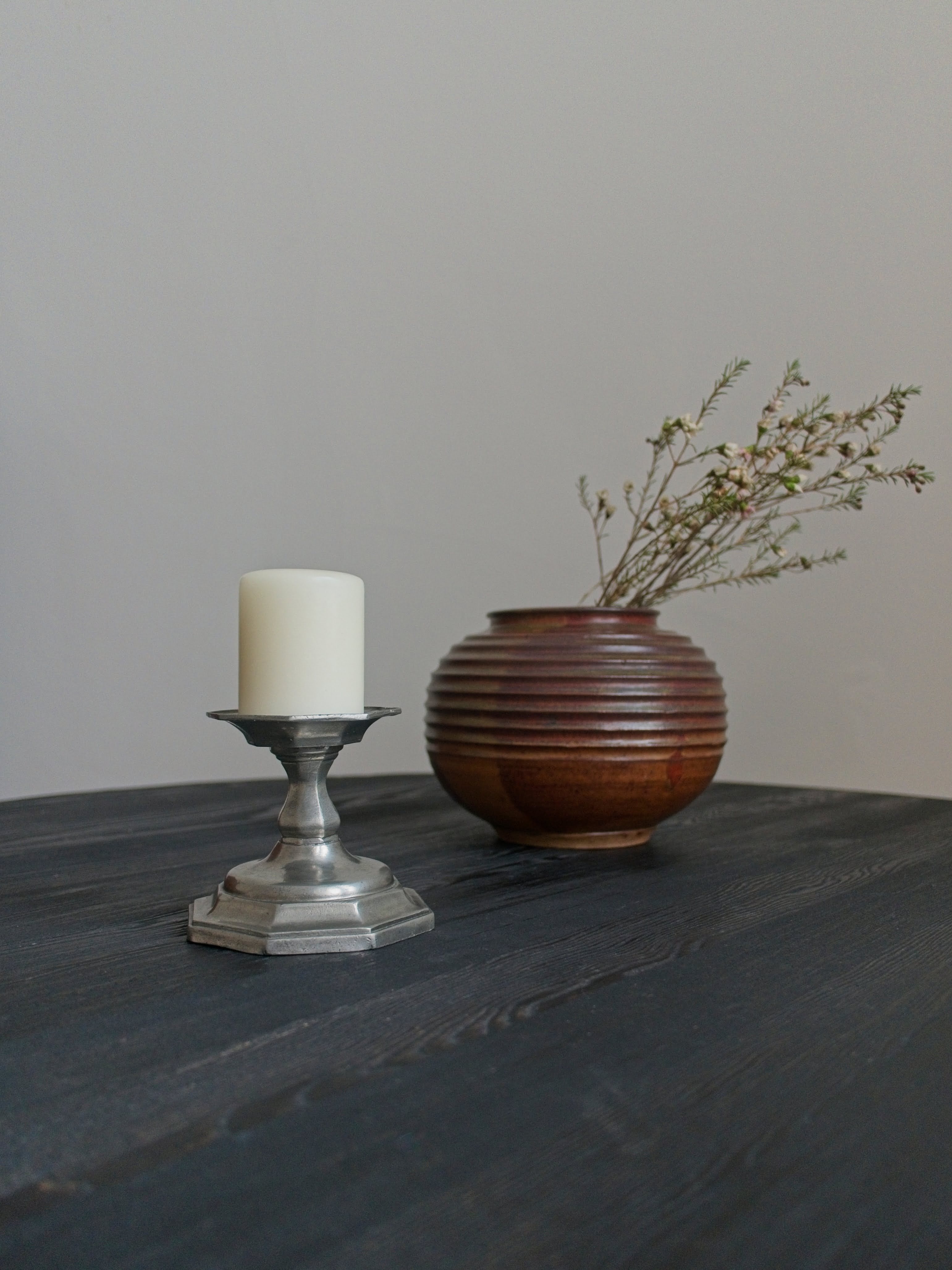 Decorative vintage pewter candle holder with cream pillar candle next to an art deco bronze globe vase with branches on a black wooden table in front of a neutral wall.