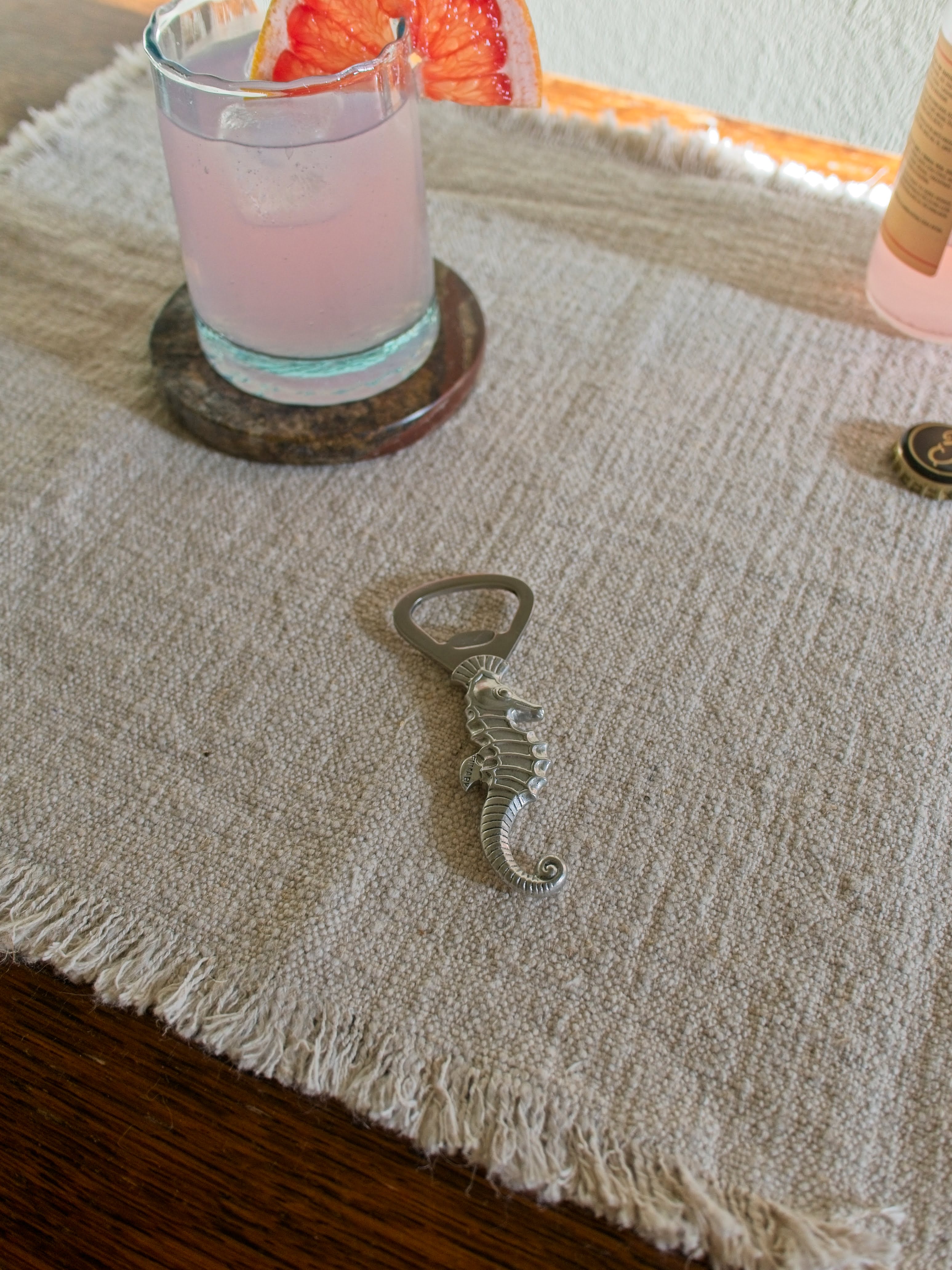 A Danish 1930s vintage art deco pewter seahorse bottle opener by Just Andersen on a textured surface with a pink cocktail in the background.