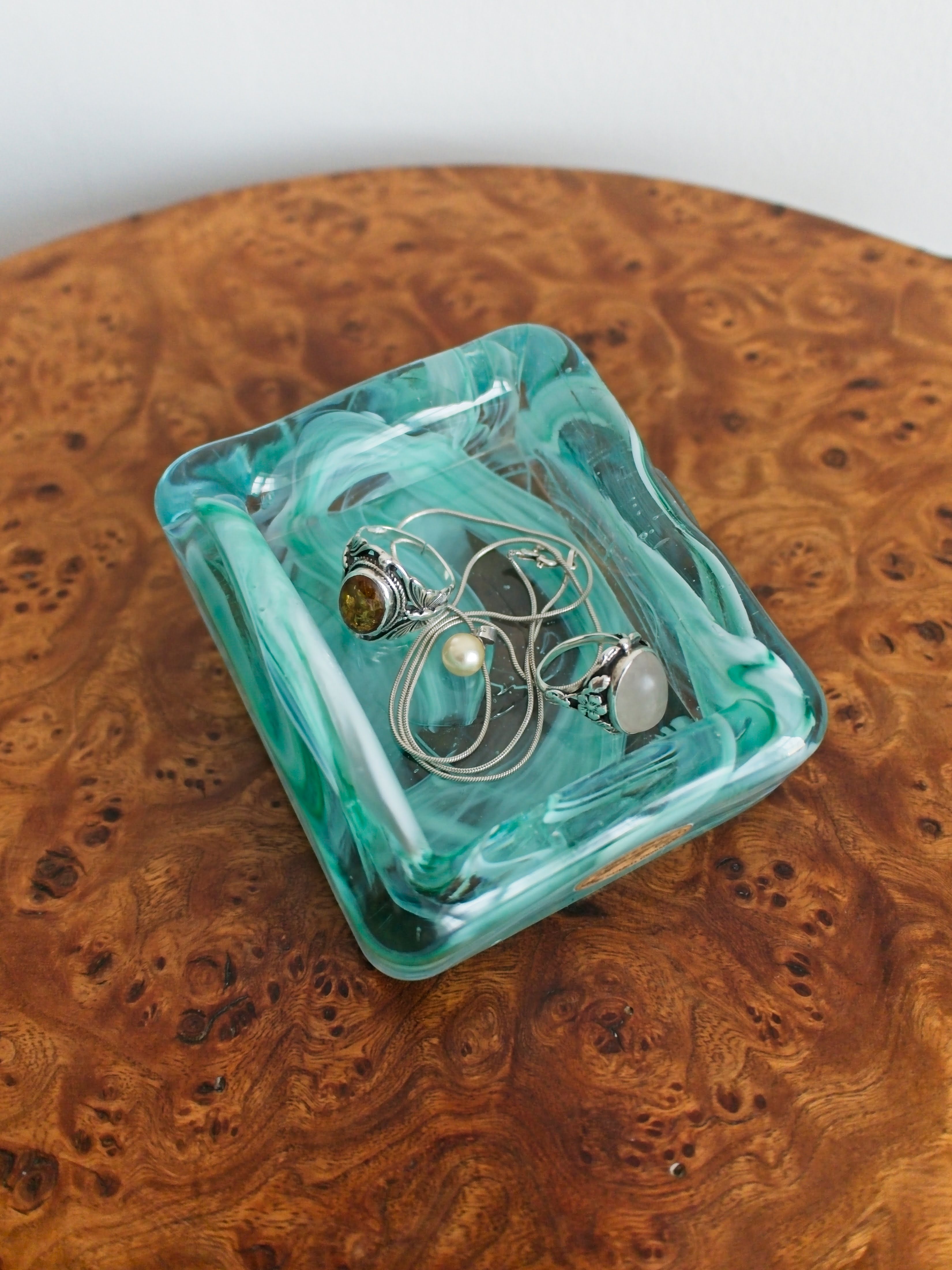 A small 1970s mid century modern glass trinket dish with green and white marble swirls similar to Murano glass containing a variety of silver jewellery set on a vintage burr wood table.