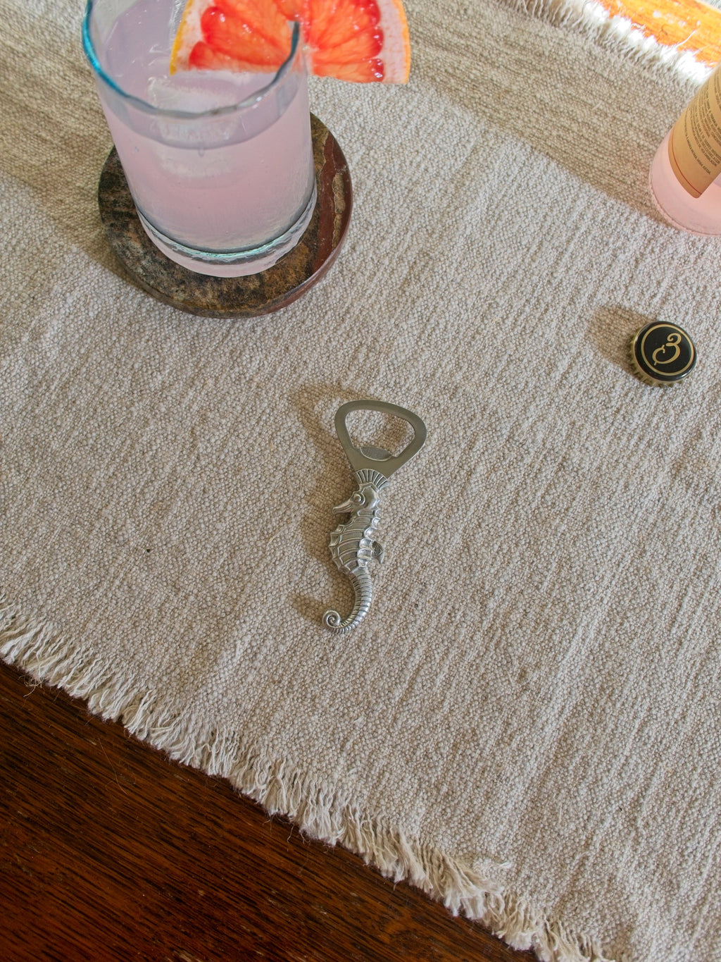 Vintage art deco pewter seahorse bottle opener by Just Andersen on a neutral linen place mat with a pink cocktail in the background.