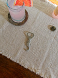 Vintage art deco pewter seahorse bottle opener by Just Andersen on a neutral linen place mat with a pink cocktail in the background.