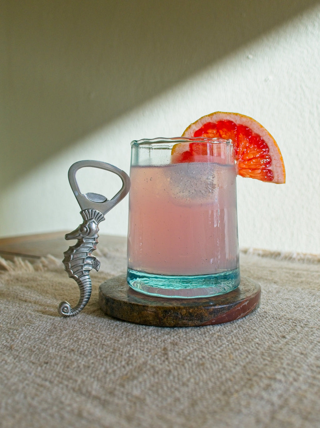 Vintage 1930s art deco pewter seahorse bottle opener by Just Andersen resting vertically on glass containing a pink cocktail with a slice of grapefruit on the side in front of a sunny green wall.