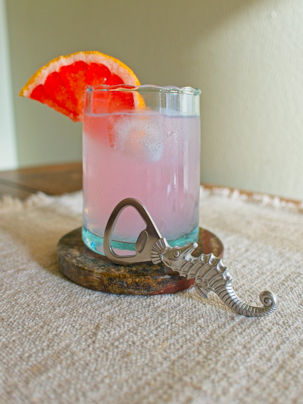 Vintage 1930s art deco pewter seahorse bottle opener by Just Andersen resting horizontally on a green marble coaster with a glass containing a pink cocktail with a slice of grapefruit on the side in front of a sunny green wall.
