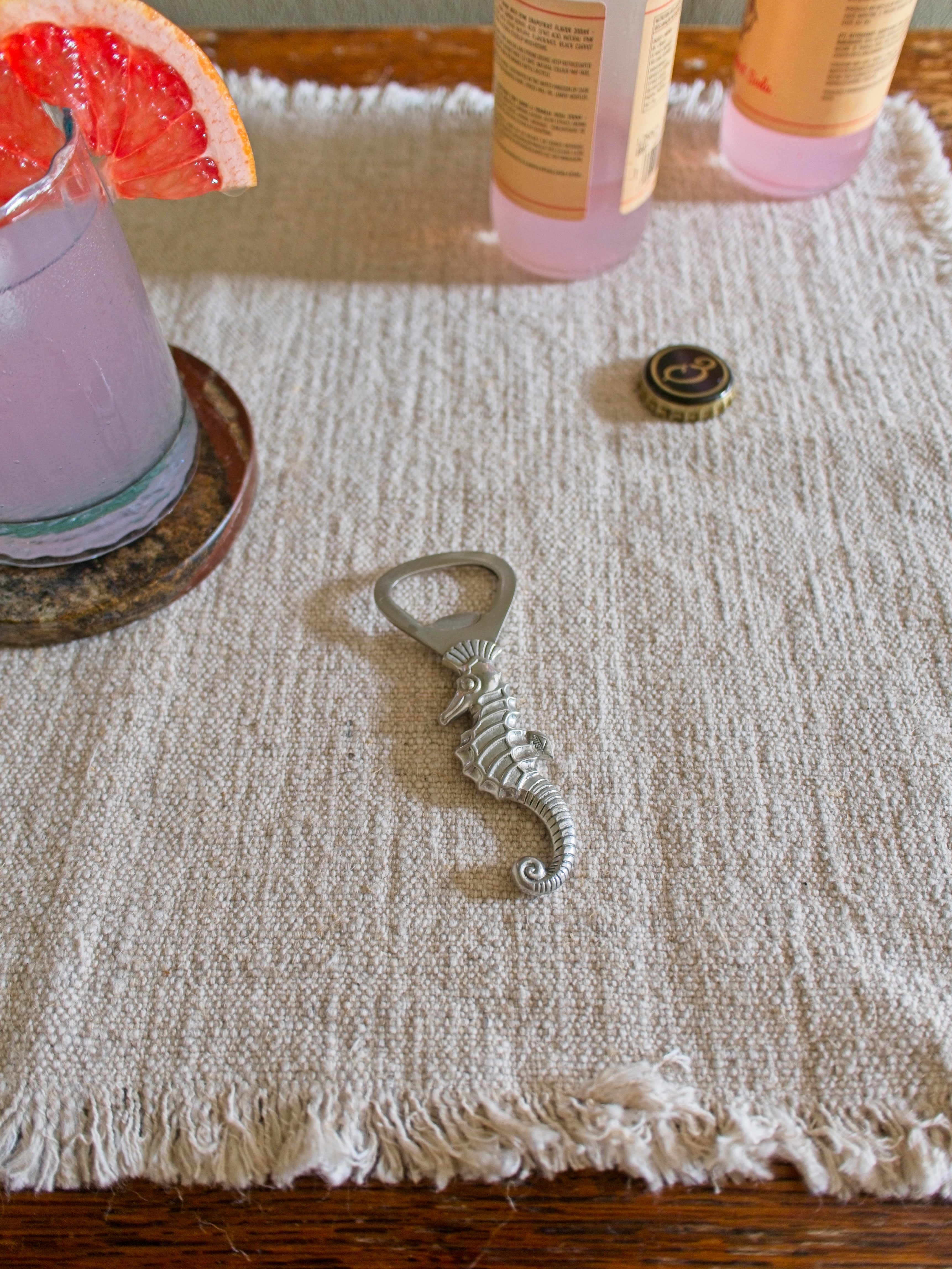 Mid century modern art deco pewter seahorse bottle opener by Just Andersen on a textured surface with a pink cocktail and bottles in the background.