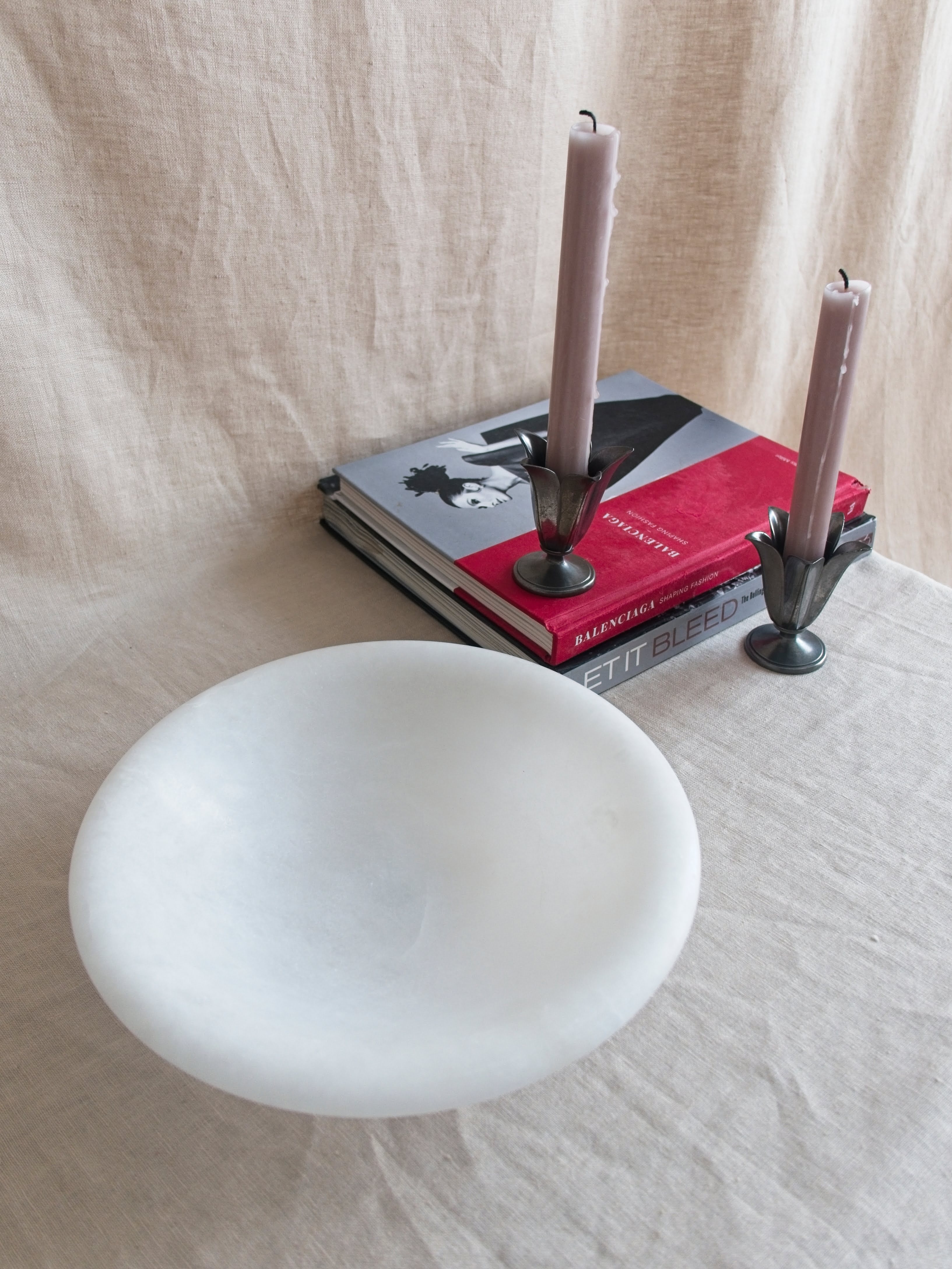 A vintage art deco style white alabaster decorative bowl by Volterra, Italy, in front of two red and grey stacked coffee table books. A pair of pewter tulip shaped vintage Just Andersen candle holders containing pink candle sticks sit on the books.