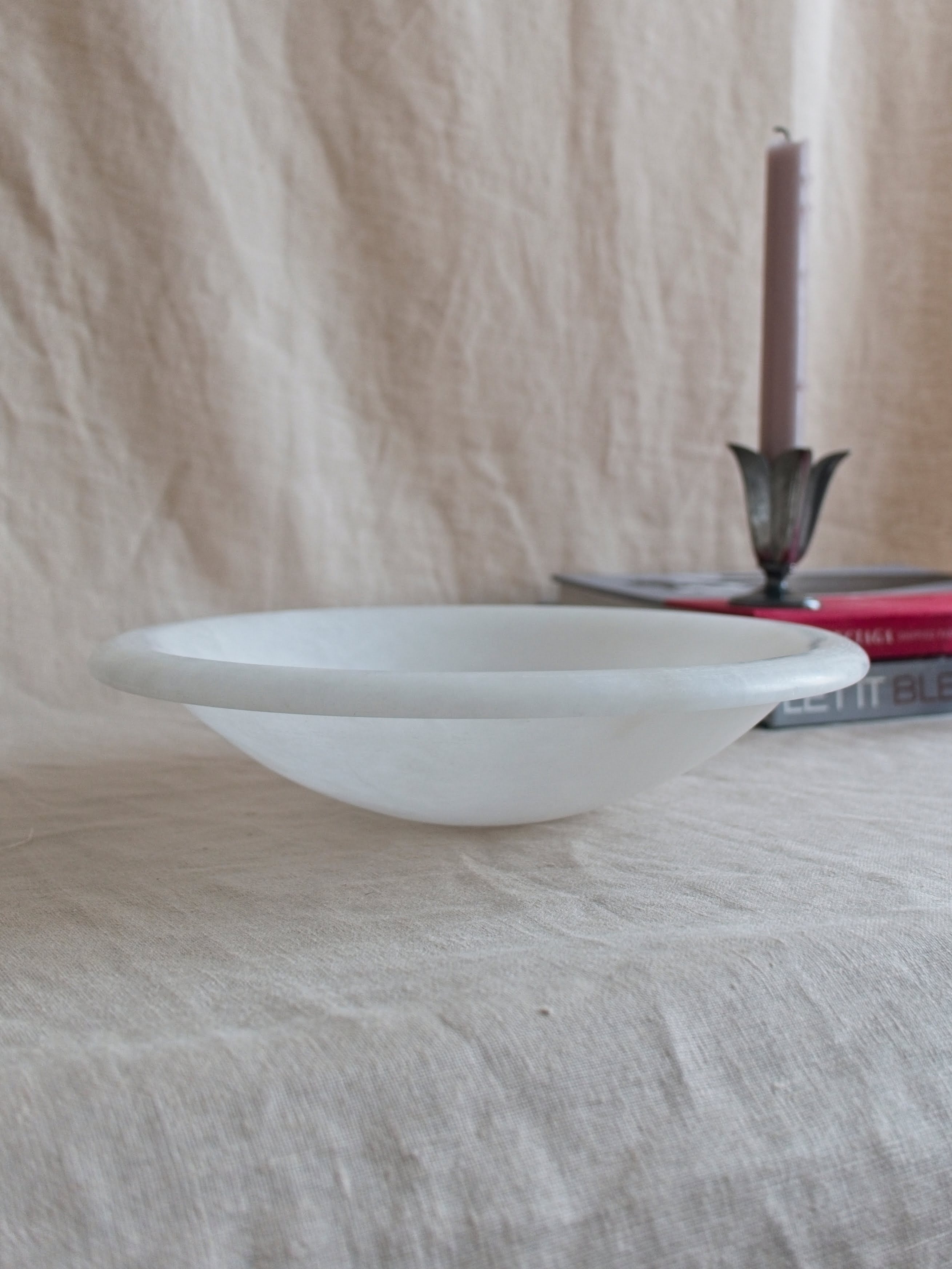 A close up of a vintage art deco style white alabaster decorative bowl by Volterra, Italy, in front of two red and grey stacked coffee table books with a pewter tulip shaped vintage Just Andersen candle holder containing a pink candle stick on a neutral linen backdrop.