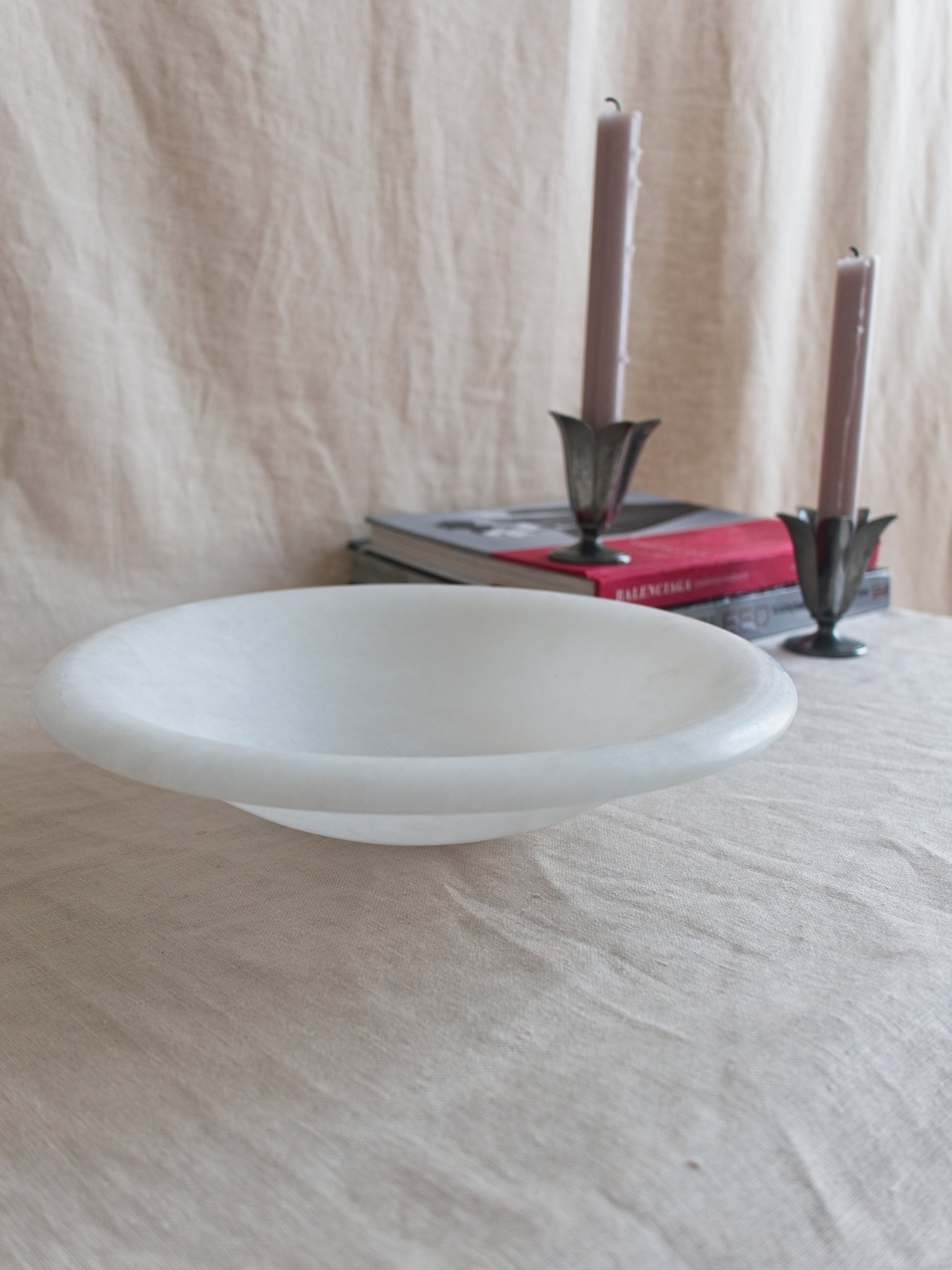 A close up on the rim of a vintage art deco style white alabaster decorative bowl by Volterra, Italy, in front of two red and grey stacked coffee table books. A pair of pewter tulip shaped vintage Just Andersen candle holders containing pink candle sticks sit on the books.