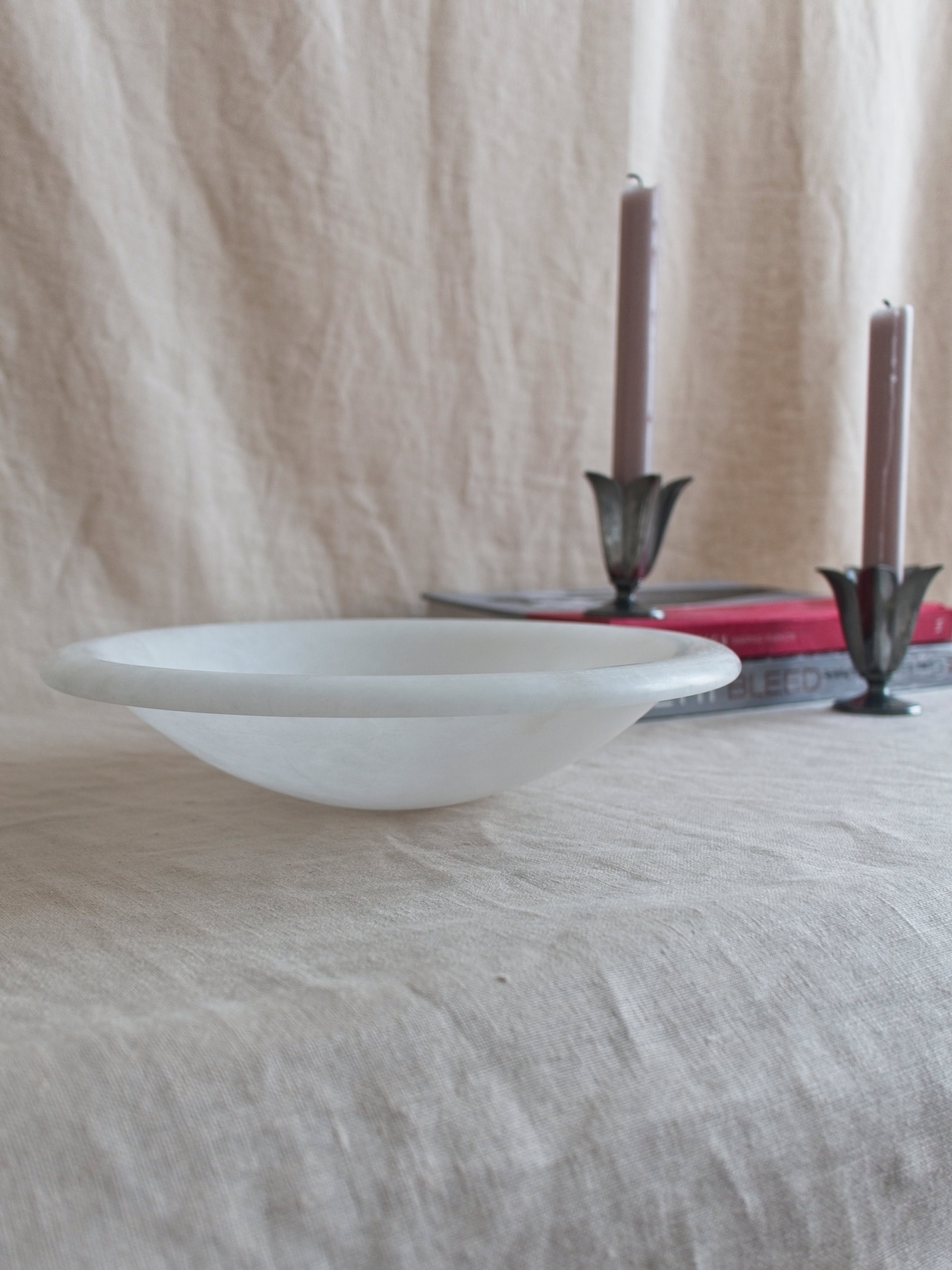 The side of a vintage art deco style white alabaster decorative bowl by Volterra, Italy, in front of two red and grey stacked coffee table books. A pair of pewter tulip shaped vintage Just Andersen candle holders containing pink candle sticks sit on the books.