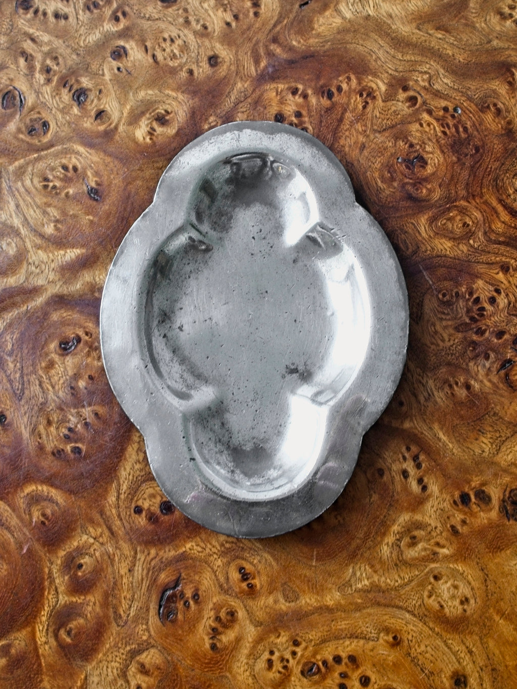 A small vintage art deco pewter ring dish by Just Andersen for GAB Sweden sitting vertically on an antique burl wood side table. The bijou floral shaped 1920s silver modernist trinket dish has four rounded petals and would make an cool and interesting addition to mid century modern, retro, baroque, modern vintage and minimalist home interiors.