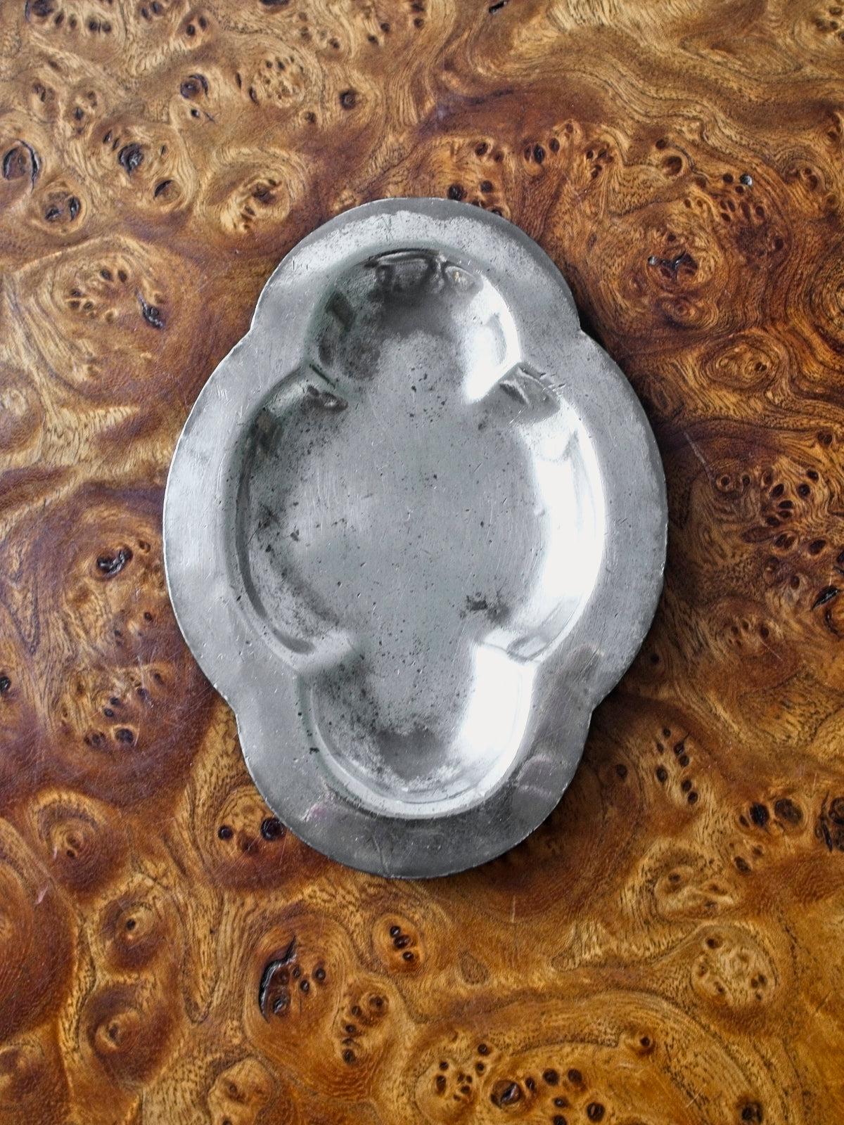 A small vintage art deco pewter ring dish by Just Andersen for GAB Sweden sitting vertically on an antique burl wood side table. The bijou floral shaped 1920s silver modernist trinket dish has four rounded petals and would make an cool and interesting addition to mid century modern, retro, baroque, modern vintage and minimalist home interiors.