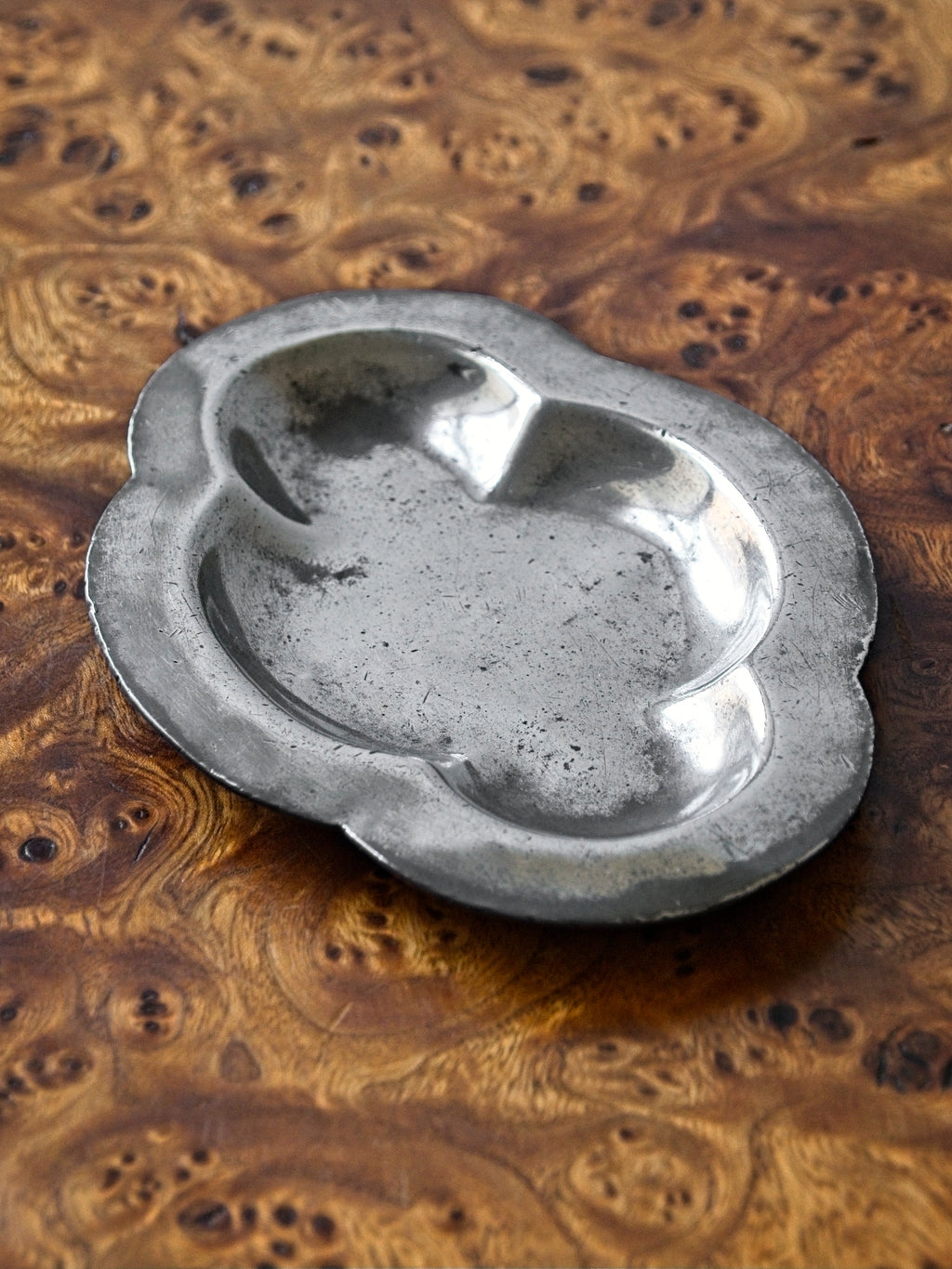 A close up of a small vintage art deco pewter ring dish by Just Andersen for GAB Sweden sitting diagonally on an antique burl wood side table. The bijou floral shaped 1920s silver modernist trinket dish has four rounded petals and would make an cool and interesting addition to mid century modern, retro, baroque, modern vintage and minimalist home interiors.