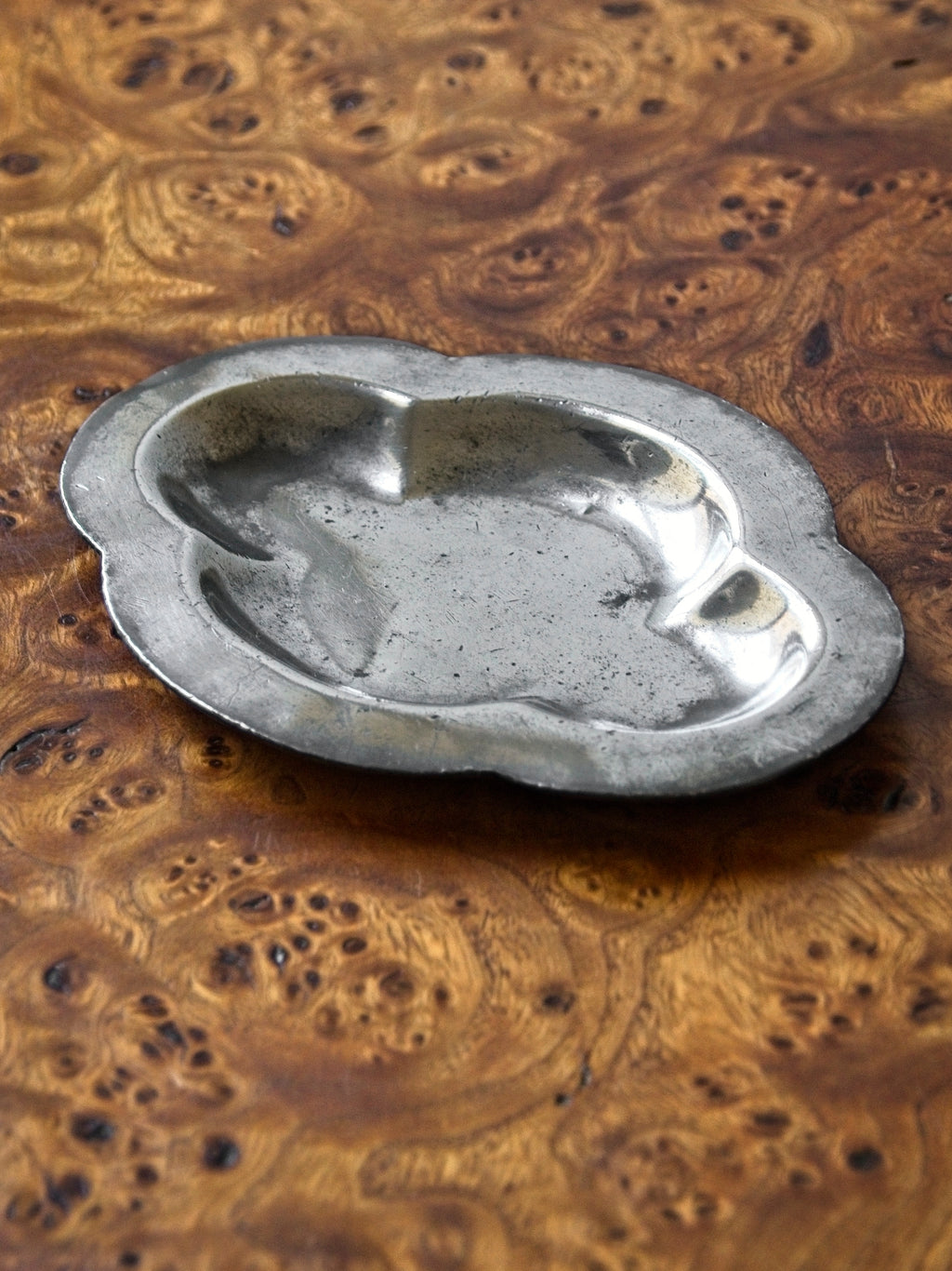 A small vintage art deco pewter ring dish by Just Andersen for GAB Sweden sitting diagonally on an antique burl wood side table. The bijou floral shaped 1920s silver modernist trinket dish has four rounded petals and would make an cool and interesting addition to mid century modern, retro, baroque, modern vintage and minimalist home interiors.