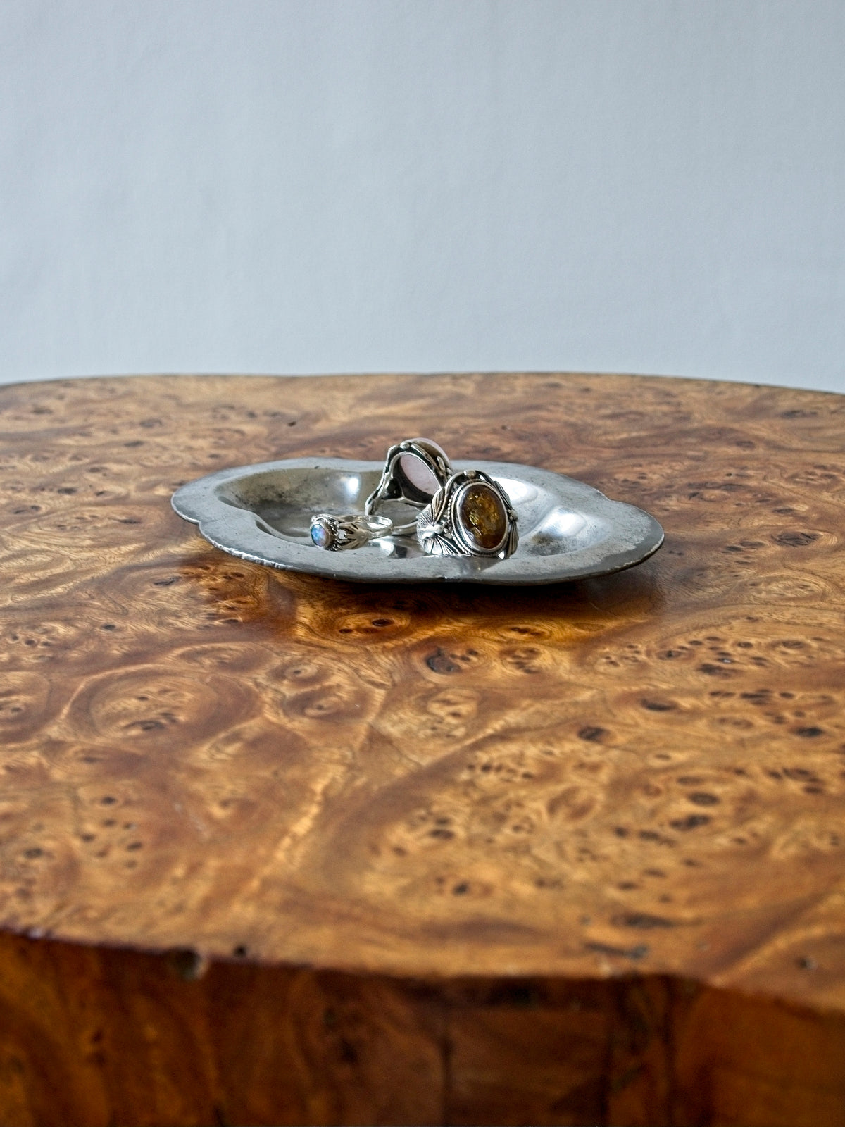A small vintage art deco pewter ring dish by Just Andersen for GAB Sweden on an antique burl wood side table in front of a white backdrop. The bijou floral shaped 1920s silver modernist trinket dish has four rounded petals is holding a selection of three stylish rings with various gems. It would make an cool and interesting addition to mid century modern, retro, baroque, modern vintage and minimalist home interiors.