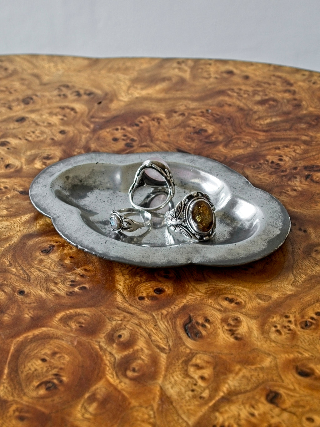 A small vintage art deco pewter ring dish by Just Andersen for GAB Sweden on an antique burl wood side table in front of a white backdrop. The bijou floral shaped 1920s silver modernist trinket dish has four rounded petals is holding a selection of three stylish rings with various gems. It would make an cool and interesting addition to mid century modern, retro, baroque, modern vintage and minimalist home interiors.