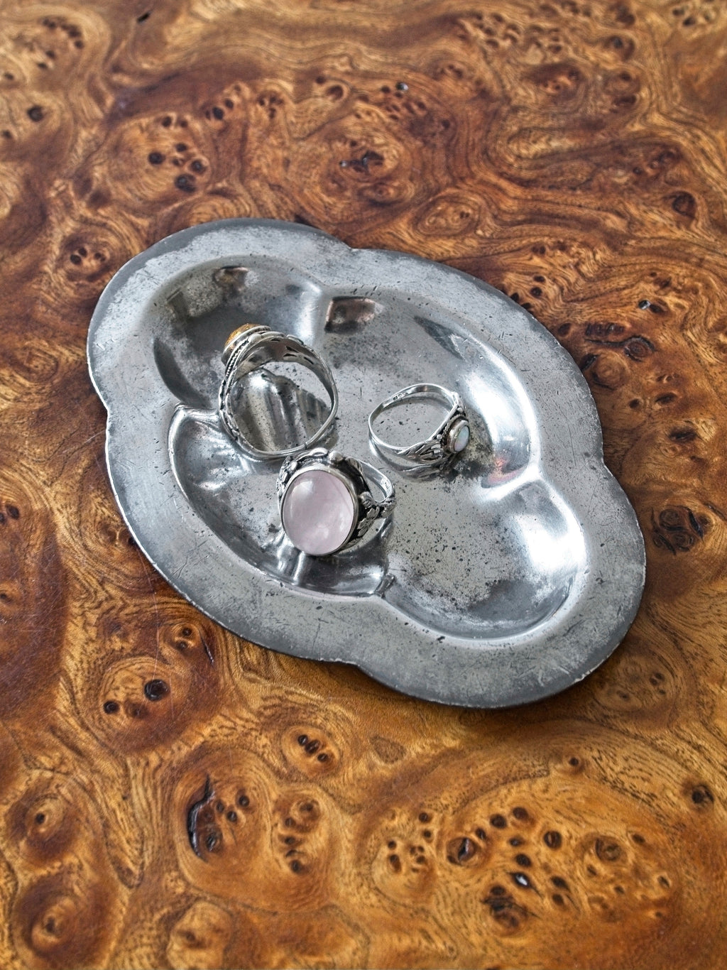 A close up of a small vintage art deco pewter ring dish by Just Andersen for GAB Sweden on an antique burl wood side table. The bijou floral shaped 1920s silver modernist trinket dish has four rounded petals is holding a selection of three stylish rings with various gems. It would make an cool and interesting addition to mid century modern, retro, baroque, modern vintage and minimalist home interiors.