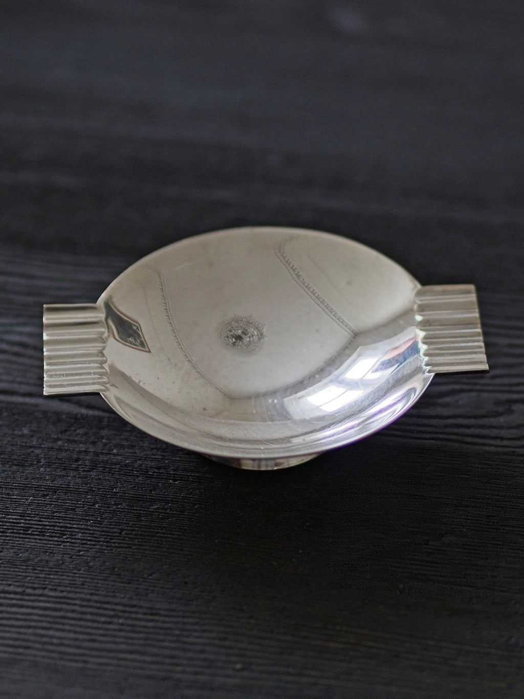 A detailed shot of a 1920s vintage modernist silver ring dish by James Dixon & Sons styled on black wood surface. Crafted from silver plate, this petite circular footed trinket dish has geometric ridged lugs and would compliment postmodern, Swedish grace, minimalist, mid century modern, Bauhaus, art deco and Hollywood Regency décor schemes. Sold by Vintrovia, vintage home décor store UK.