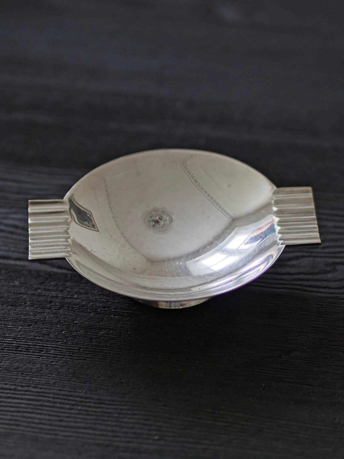 A detailed shot of a 1920s vintage modernist silver ring dish by James Dixon & Sons styled on black wood surface. Crafted from silver plate, this petite circular footed trinket dish has geometric ridged lugs and would compliment postmodern, Swedish grace, minimalist, mid century modern, Bauhaus, art deco and Hollywood Regency décor schemes. Sold by Vintrovia, vintage home décor store UK.