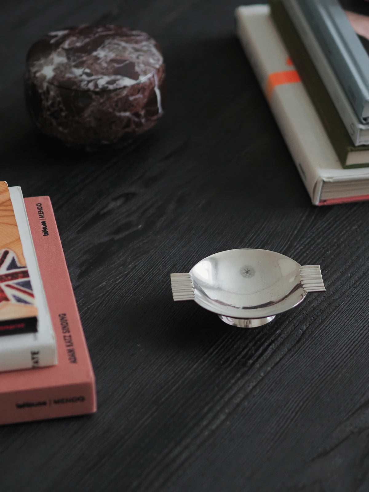 A 1920s vintage modernist silver ring dish by James Dixon & Sons styled black with a red marble box and two stacks of stylish books in the background. Crafted from silver plate, this petite circular footed trinket dish with geometric ridged lugs would compliment postmodern, art deco, minimalist, mid century modern, Bauhaus, Swedish grace and Hollywood Regency décor schemes. Sold by Vintrovia, vintage home décor store UK.