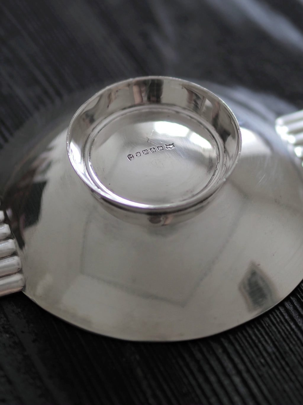 A close up of the makers mark on the base of a 1920s vintage modernist silver ring dish by James Dixon & Sons styled on black wood surface. Crafted from silver plate, this petite circular footed trinket dish has geometric ridged lugs and would compliment postmodern, Swedish grace, minimalist, mid century modern, Bauhaus, art deco and Hollywood Regency décor schemes. Sold by Vintrovia, vintage home décor store UK.