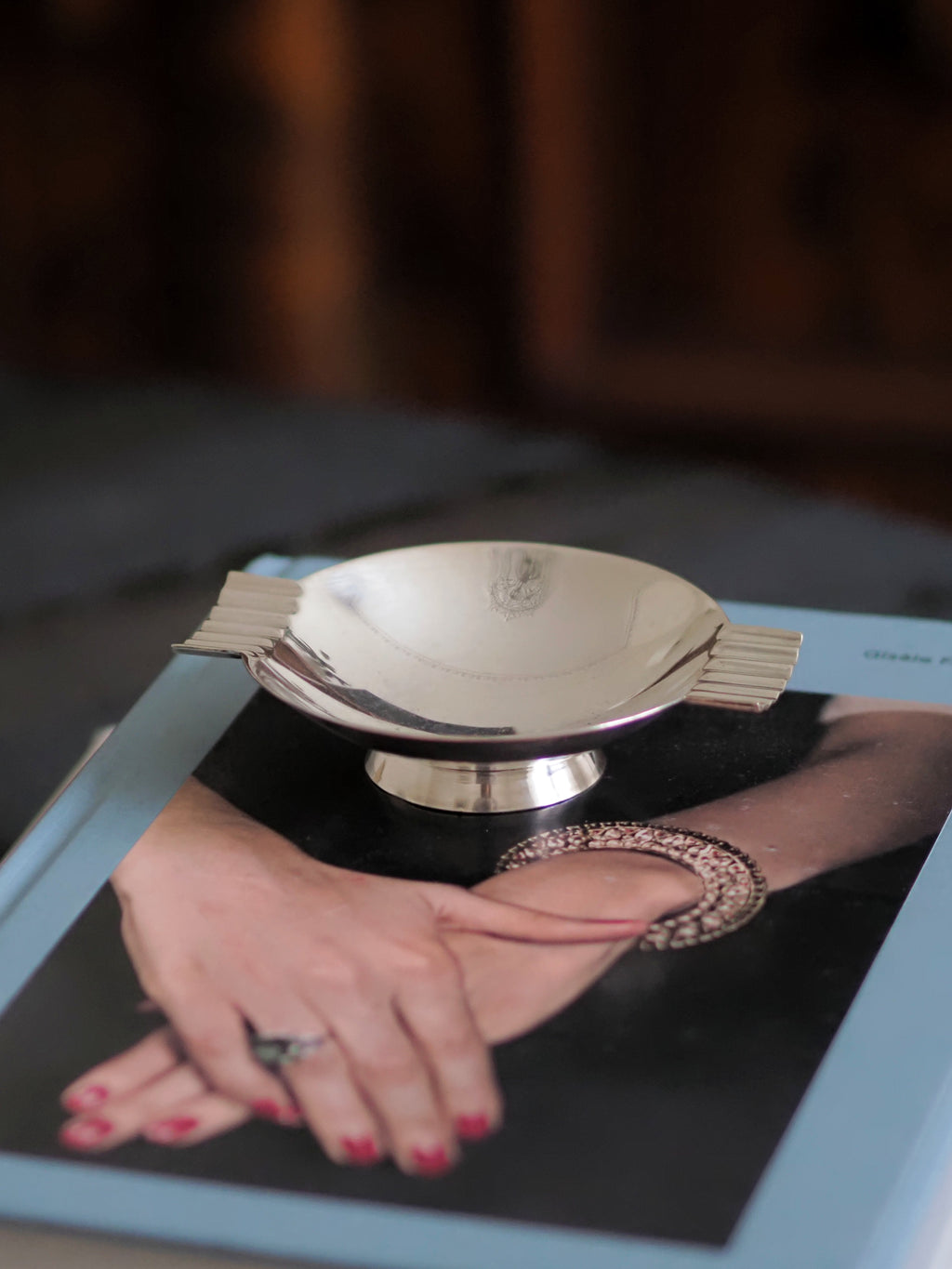 A vintage modernist silver ring dish by James Dixon & Sons styled on a stack of books on a black coffee table with a walnut sideboard in the background. Crafted from silver plate, this petite circular geometric footed trinket dish would compliment postmodern, Swedish grace, minimalist, mid century modern, Bauhaus, art deco and Hollywood Regency décor schemes. Sold by Vintrovia, vintage home décor store UK.