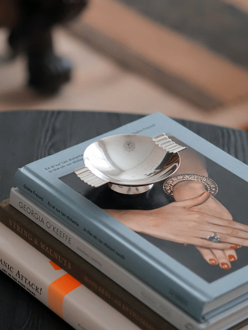 A vintage modernist silver ring dish by James Dixon & Sons styled on a stack of books on a black coffee table with a striped rug in the background. Crafted from silver plate, this petite circular geometric footed trinket dish would compliment postmodern, Swedish grace, minimalist, mid century modern, Bauhaus, art deco and Hollywood Regency décor schemes. Sold by Vintrovia, vintage home décor store UK.