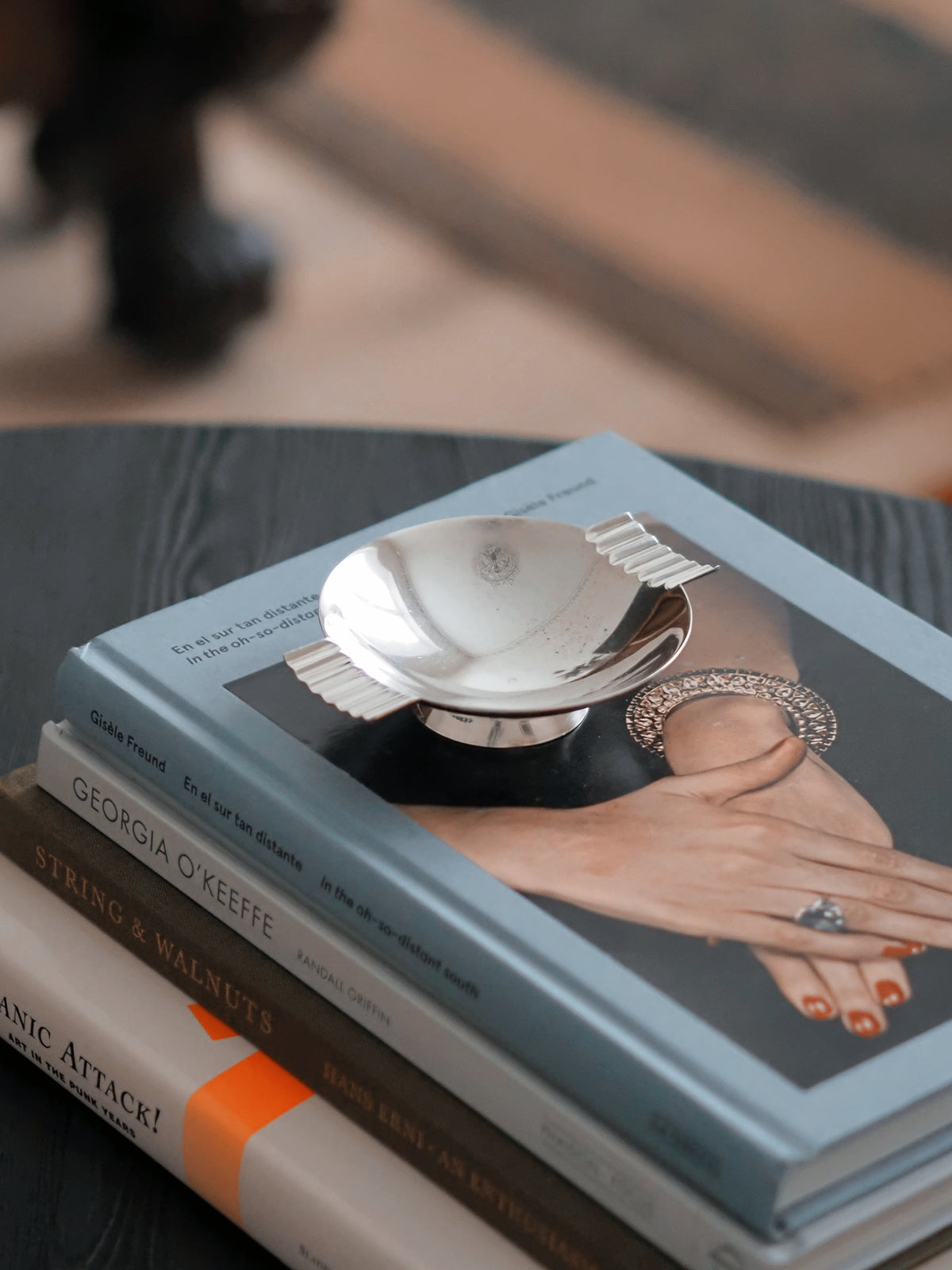 A vintage modernist silver ring dish by James Dixon & Sons styled on a stack of books on a black coffee table with a striped rug in the background. Crafted from silver plate, this petite circular geometric footed trinket dish would compliment postmodern, Swedish grace, minimalist, mid century modern, Bauhaus, art deco and Hollywood Regency décor schemes. Sold by Vintrovia, vintage home décor store UK.