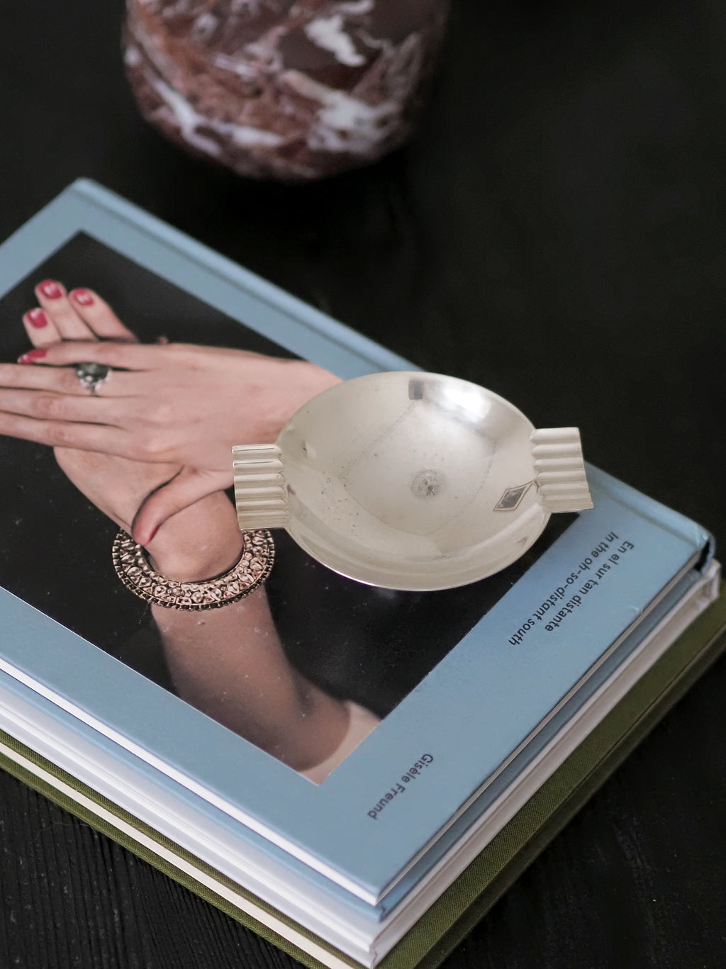 A vintage modernist silver ring dish by James Dixon & Sons styled on a stack of books on a black coffee table with a red marble box in the background. Crafted from silver plate, this petite circular geometric footed trinket dish would compliment postmodern, Swedish grace, minimalist, mid century modern, Bauhaus, art deco and Hollywood Regency décor schemes. Sold by Vintrovia, vintage home décor store UK.