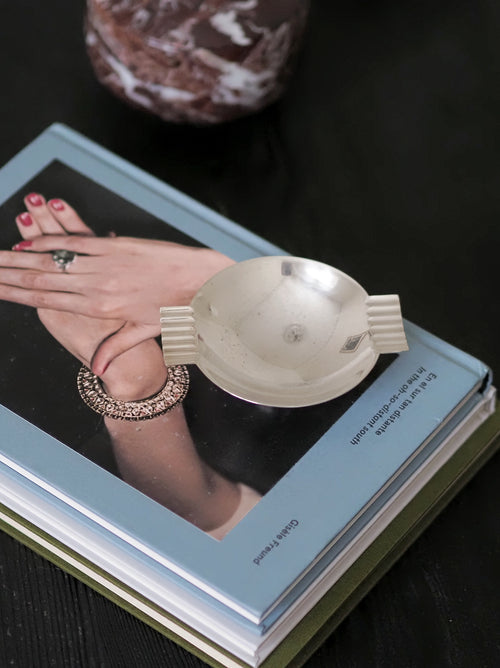 A vintage modernist silver ring dish by James Dixon & Sons styled on a stack of books on a black coffee table with a red marble box in the background. Crafted from silver plate, this petite circular geometric footed trinket dish would compliment postmodern, Swedish grace, minimalist, mid century modern, Bauhaus, art deco and Hollywood Regency décor schemes. Sold by Vintrovia, vintage home décor store UK.