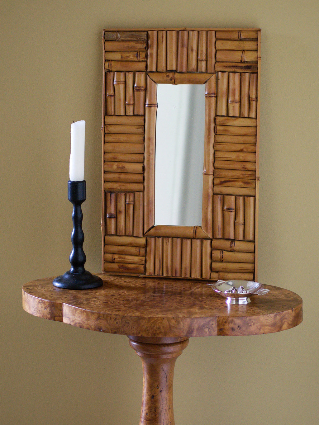 A vintage 1970s wall mounted bamboo mirror by UK modern vintage home décor store Vintrovia resting on an antique burl wood side table with a vintage bobbin wood candle holder and a silver modernist ring dish in front of a yellow wall. This mid century modern mirror is hand crafted using individual honey-toned rattan pieces to form a geometric check design that creates an interesting, modernist aesthetic. A versatile retro wall décor piece that would elevate boho, eclectic, coastal and rustic home interiors.