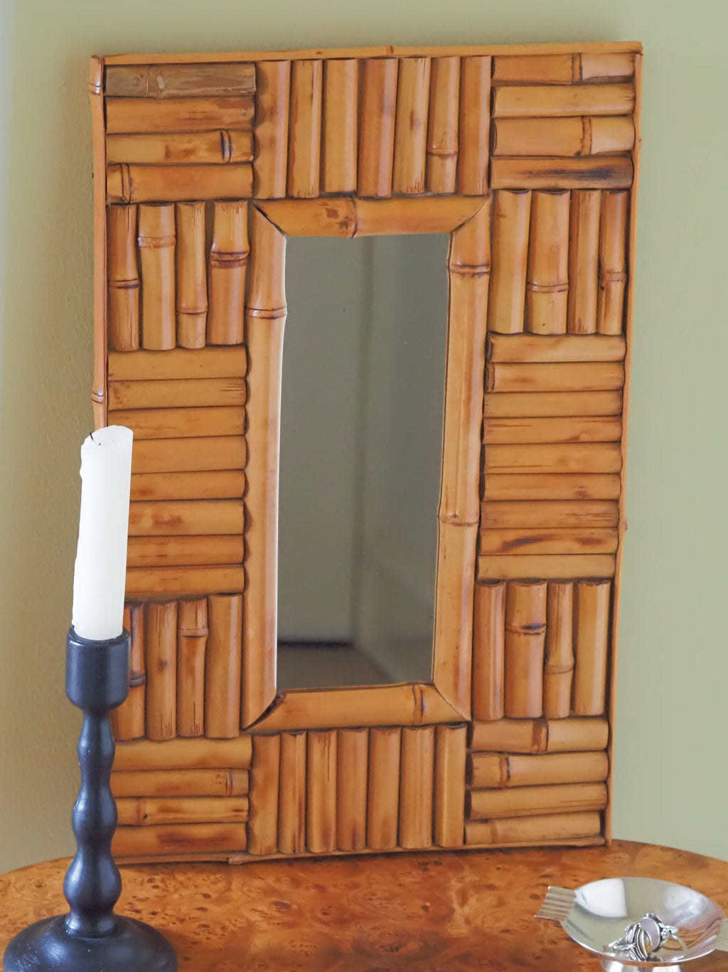A vintage 1970s wall mounted bamboo mirror by UK modern vintage home décor store Vintrovia resting on an antique burl wood side table with a vintage bobbin wood candle holder and a silver modernist ring dish in front of a yellow wall. This mid century modern mirror is hand crafted using individual honey-toned rattan pieces to form a geometric check design that creates an interesting, modernist aesthetic. A versatile retro wall décor piece that would elevate boho, eclectic, coastal and rustic home interiors.