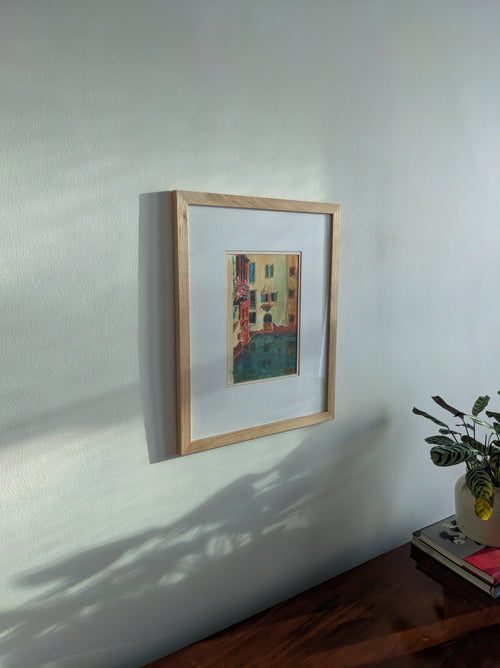 Framed vintage watercolour painting of a colourful Venetian canal scene on a white wall with a plant to the right.