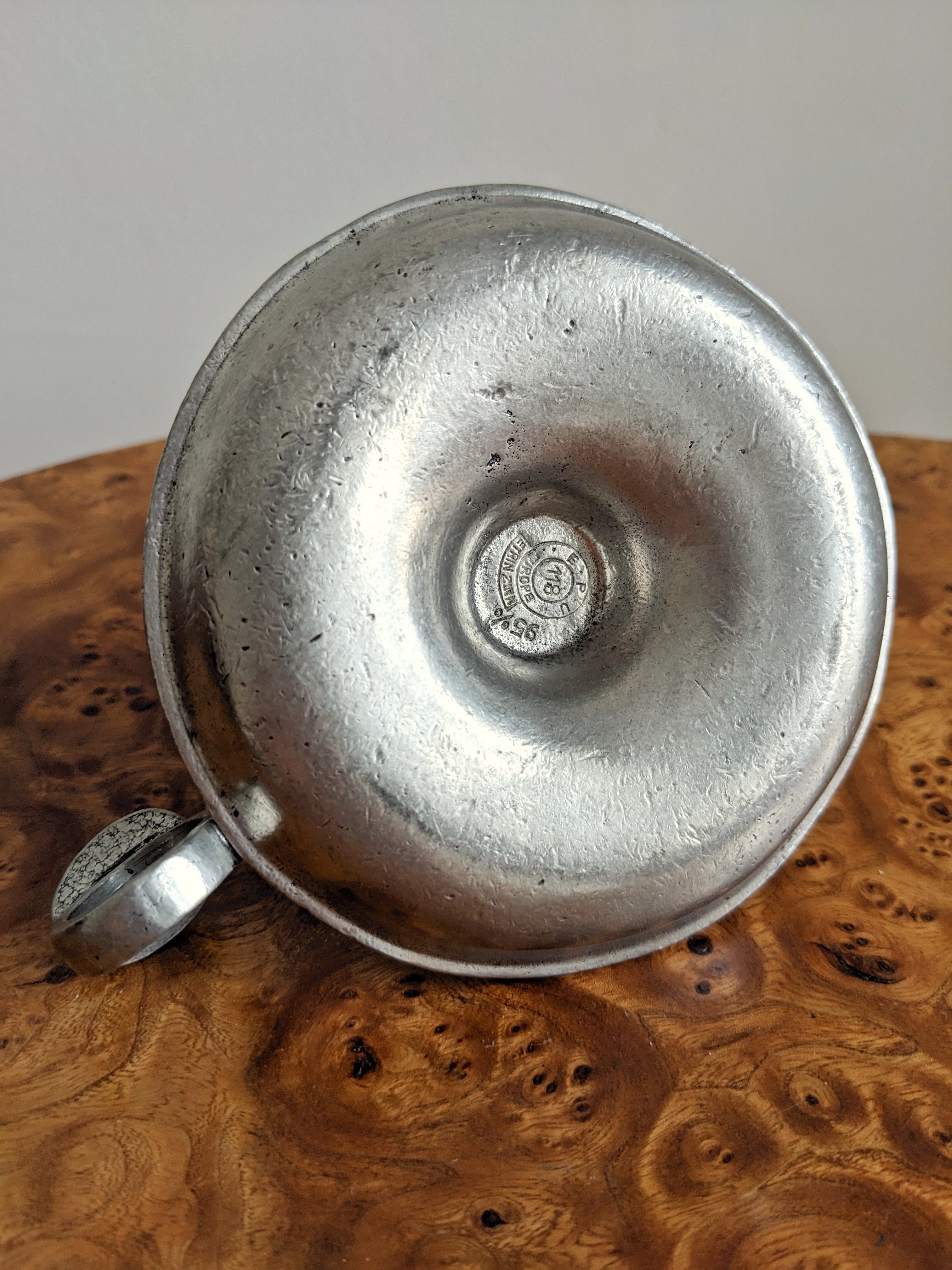 A close up of the bottom of a vintage circular silver candle holder showing a pewter stamp that says 'E.P.U. Europe, Etain Zinn'. the Candle holder sits on a burr walnut side table in front of a white background.