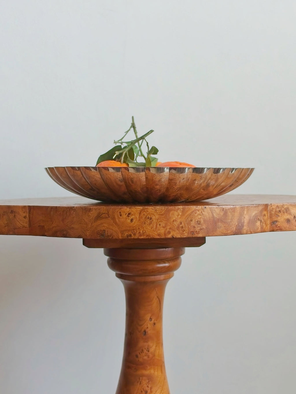 A vintage silver shell shaped bowl containing oranges on an antique burr wood table against a plain background.
