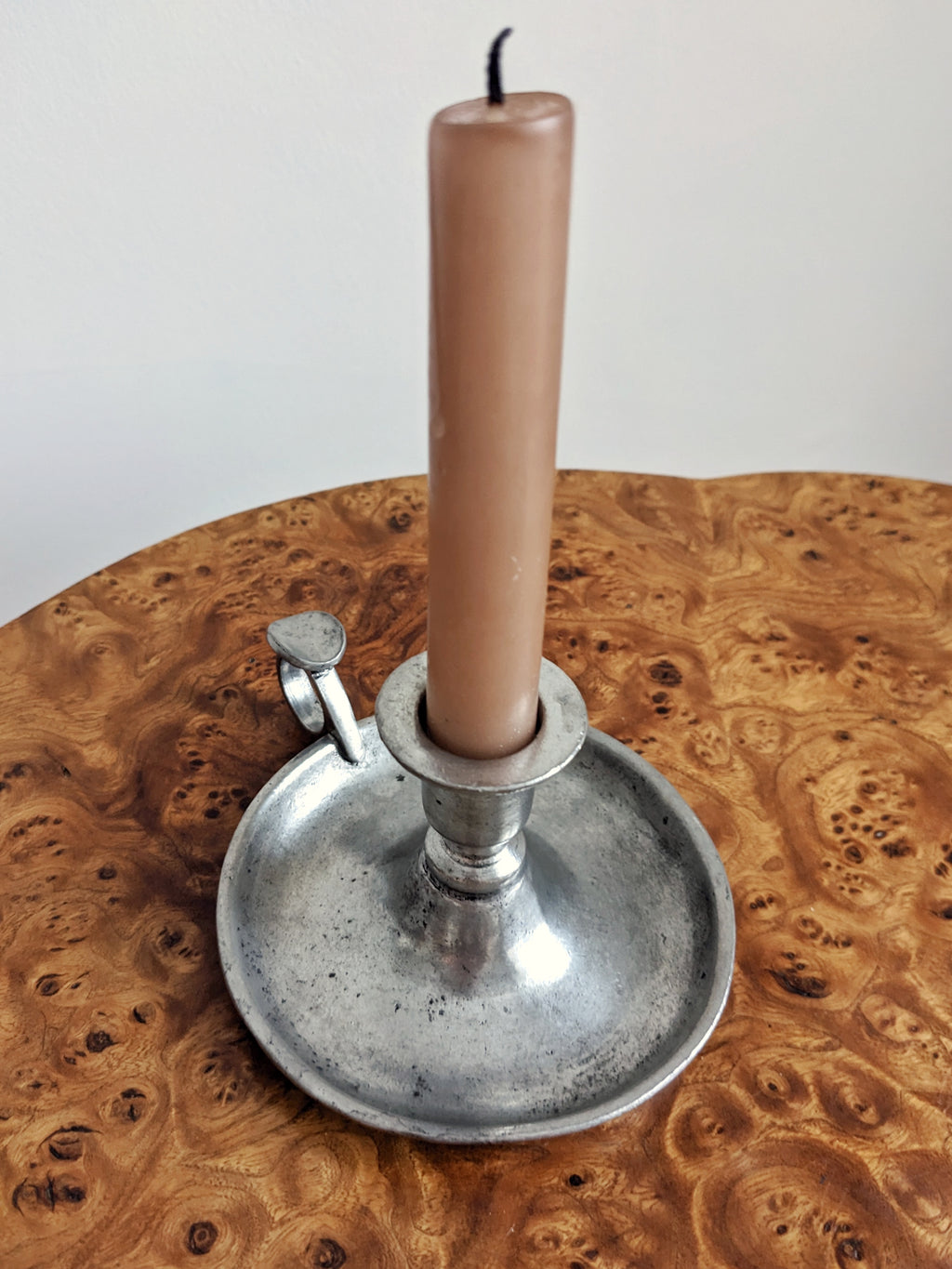A close up of a pink candle in an antique style silver candle holder on a burr wood table with a white background.