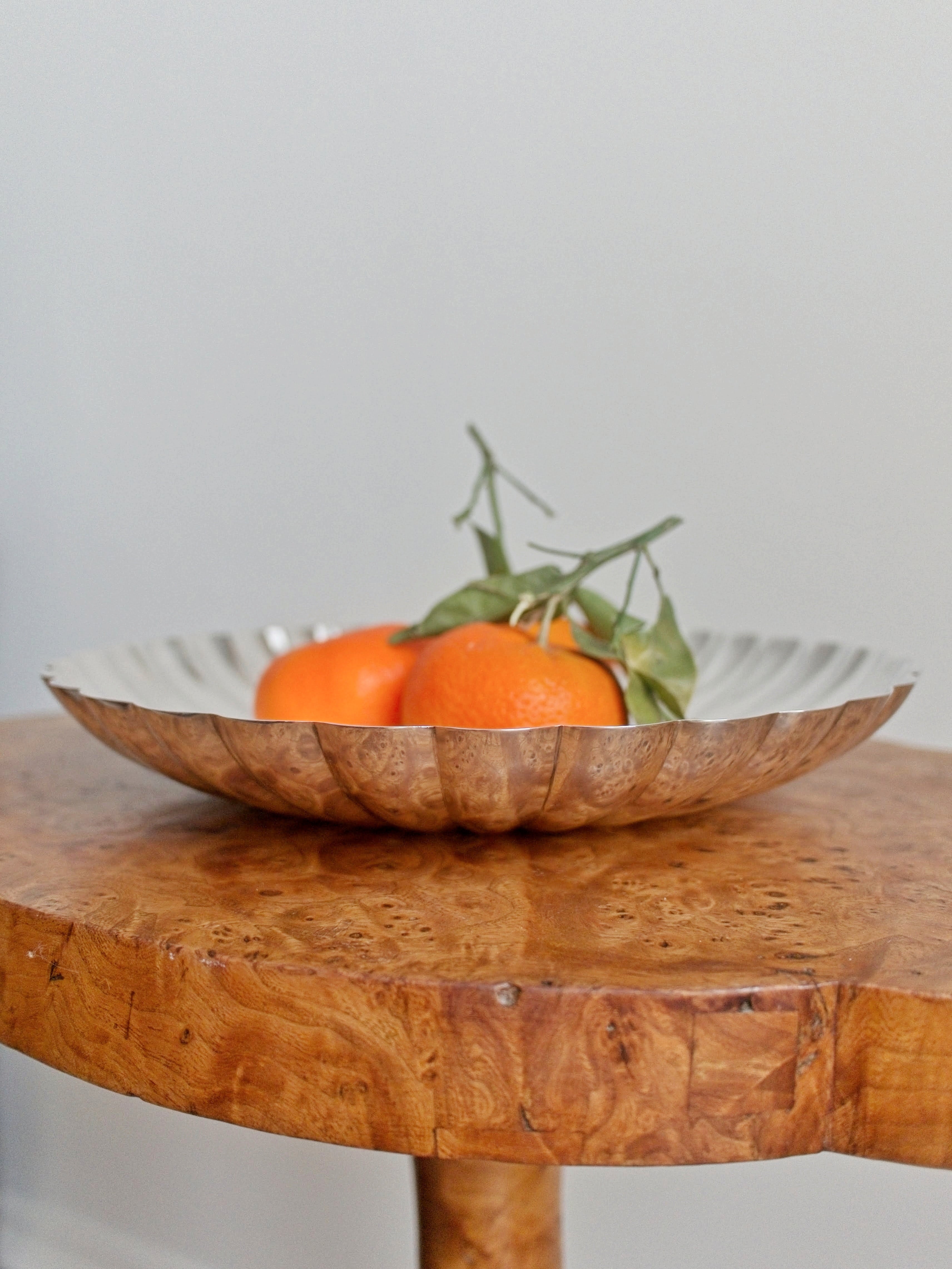 A close up of the side of a vintage scalloped Elkington & Co silver bowl with three oranges inside sitting on an antique burr walnut side table n front of a neutral background.
