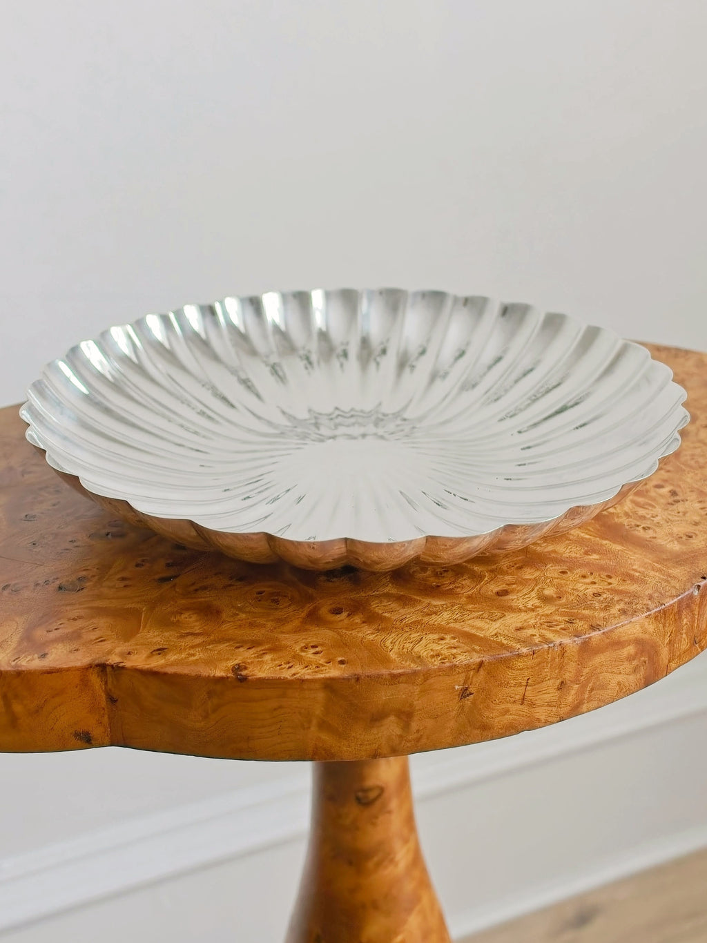 An antique burr wood side table with a vintage shell shaped silver decorative bowl in front of a plain background.