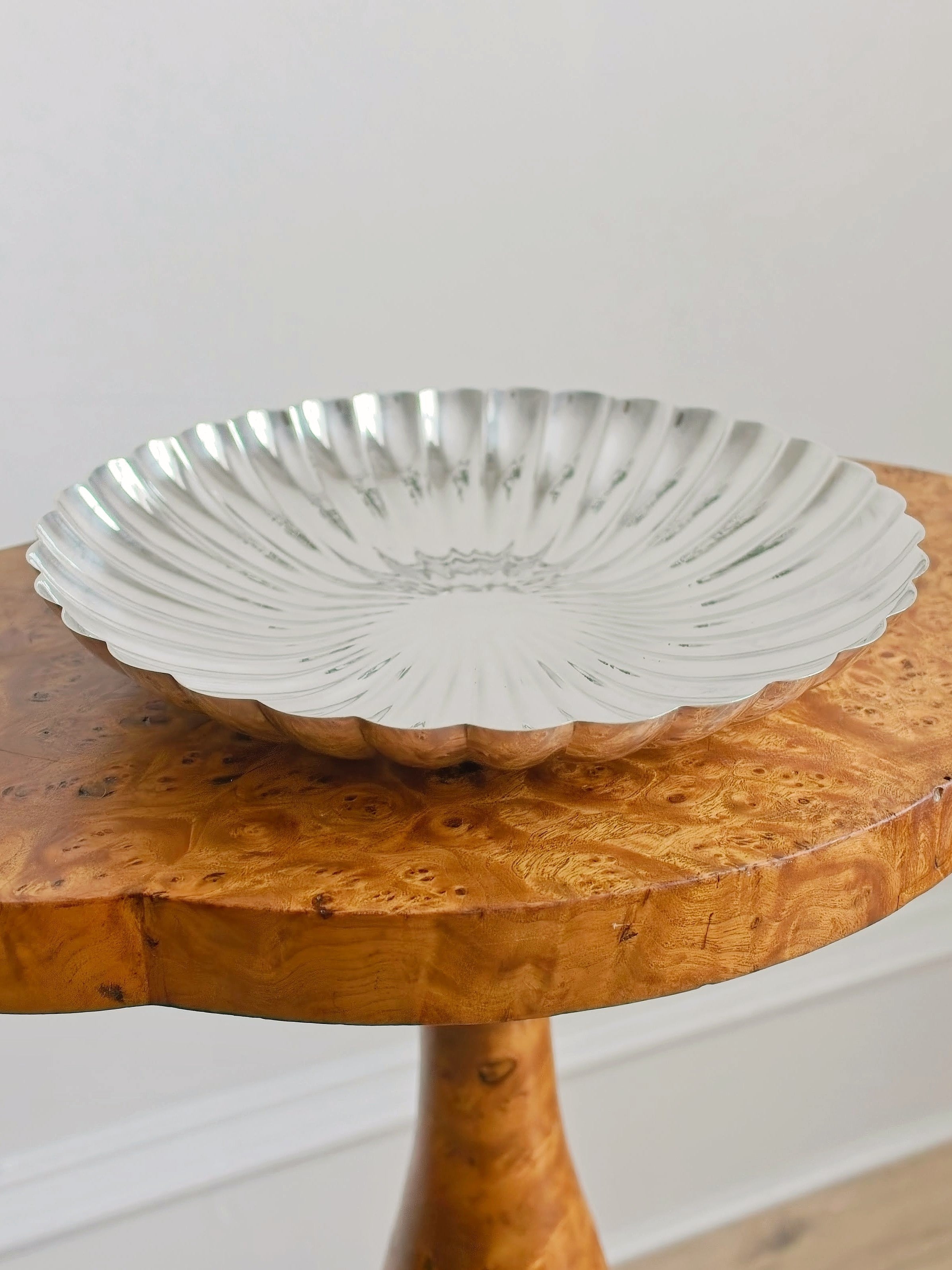 An antique burr wood side table with a vintage shell shaped silver decorative bowl in front of a plain background.