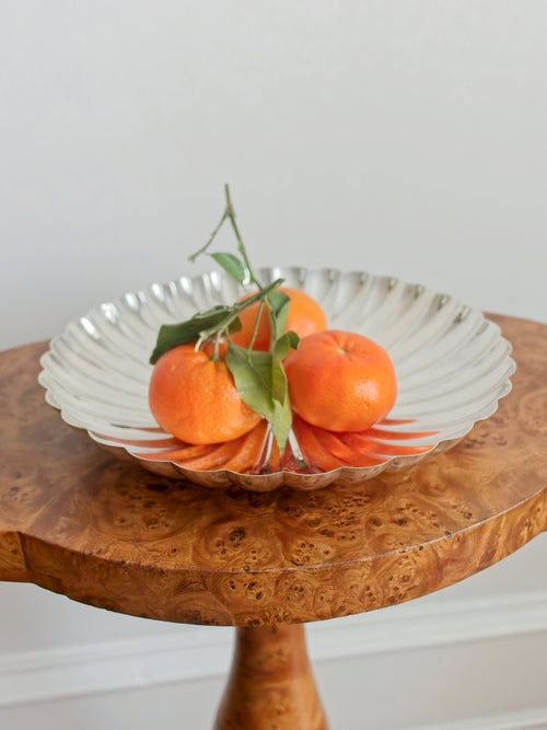 A vintage silver scallop shaped bowl containing three oranges on a Georgian burr wood table against a plain wall.