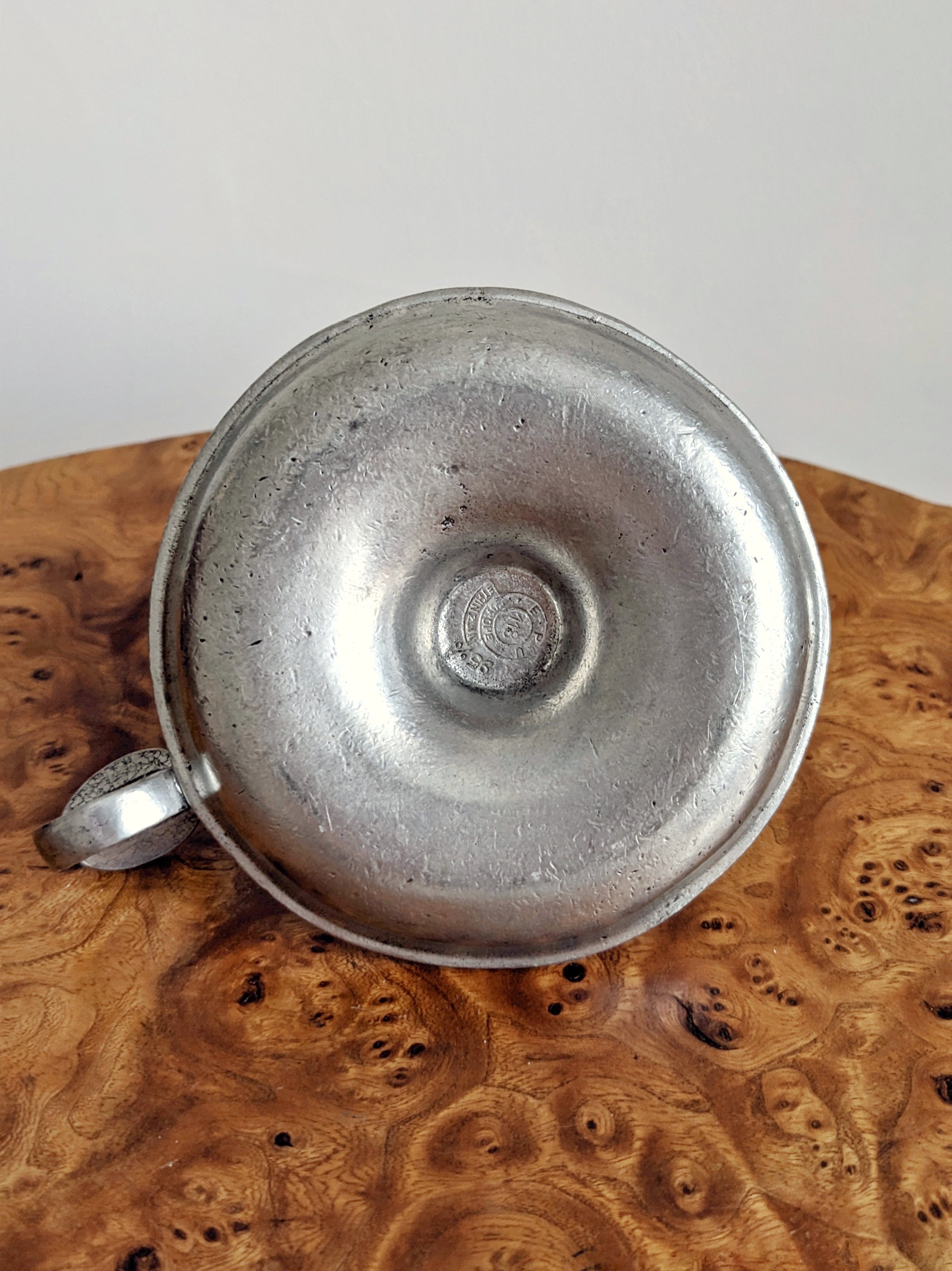 A close up of the bottom of a vintage circular silver candle holder showing a pewter stamp that says 'E.P.U. Europe, Etain Zinn'. The candle holder sits on a Georgian burr walnut side table in front of a white background.