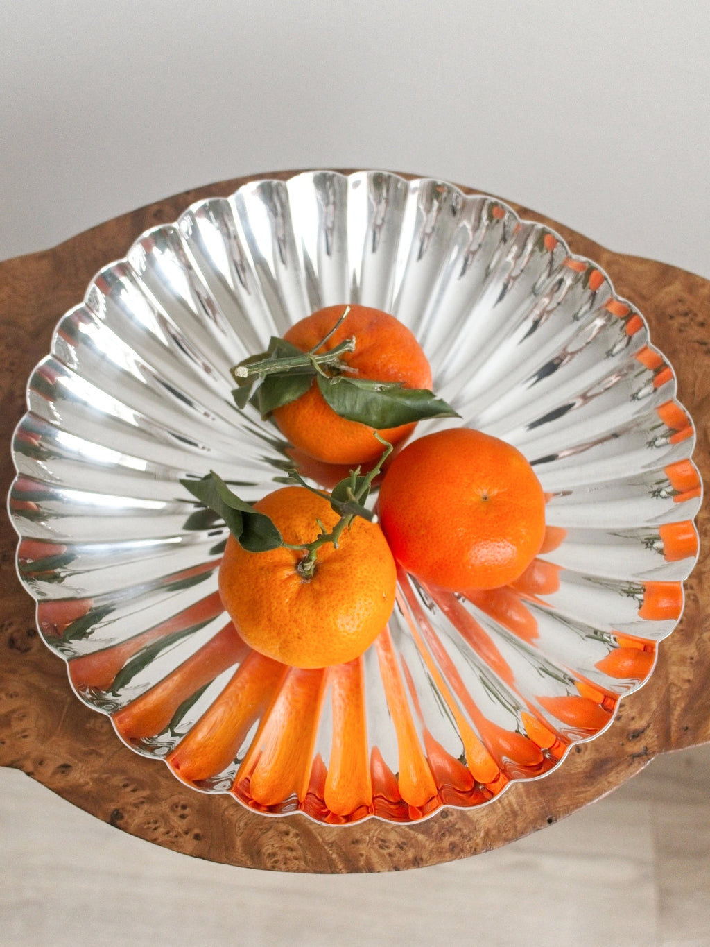 A birds eye view of a vintage scallop edged silver bowl containing three oranges with stems and leaves attached on a brown walnut burr wood side table.